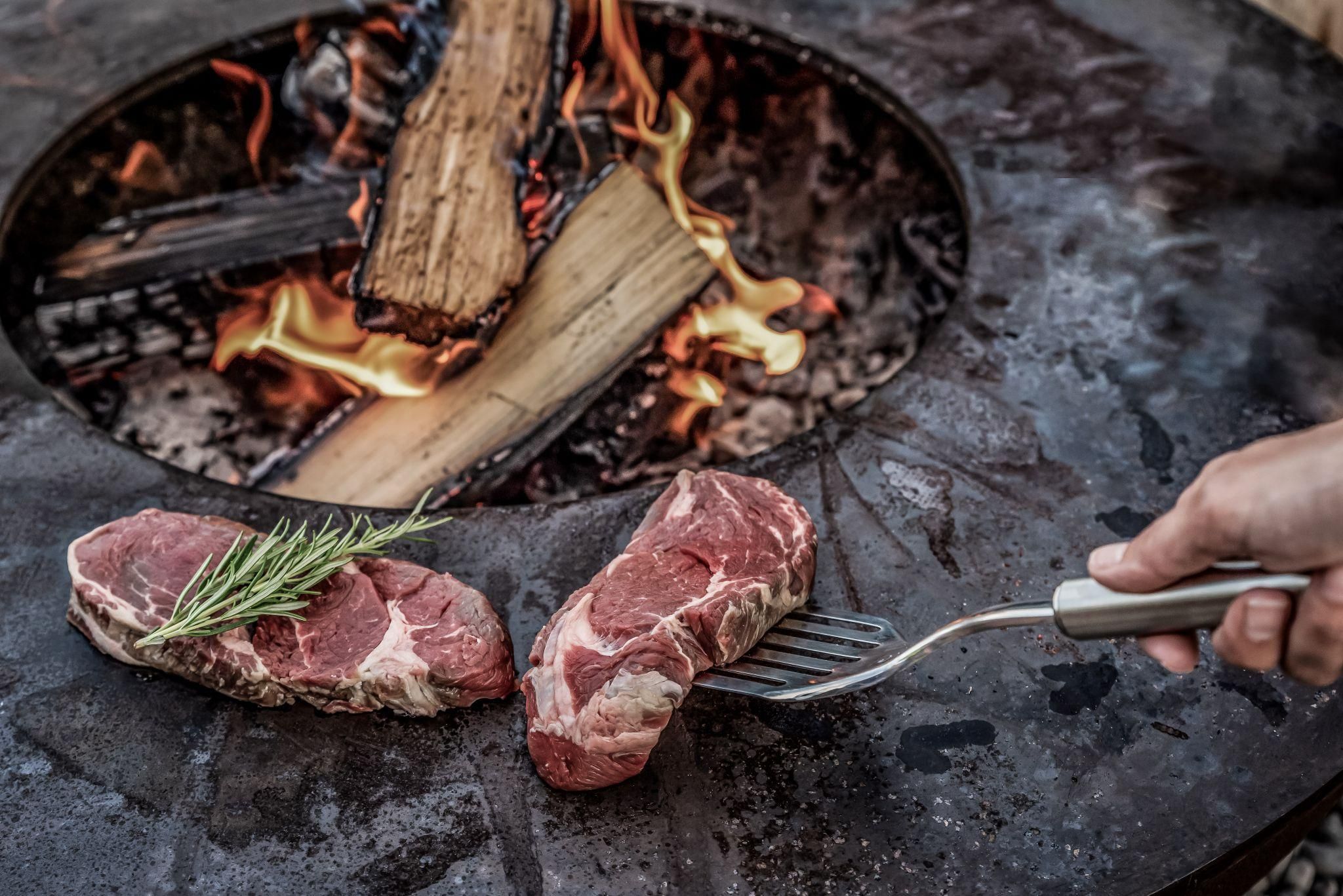 Rohes Fleisch liegt auf einem Grill, während es über offenem Feuer gart. Im Hintergrund brennen Holzscheite und erzeugen eine gemütliche Atmosphäre.
