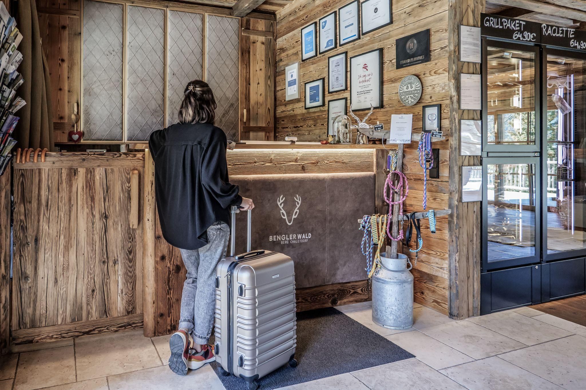 Eine Person steht an der Rezeption vom Chalet Dorf mit einem Koffer. An der Wand hängen verschiedene Auszeichnungen und Plakate.