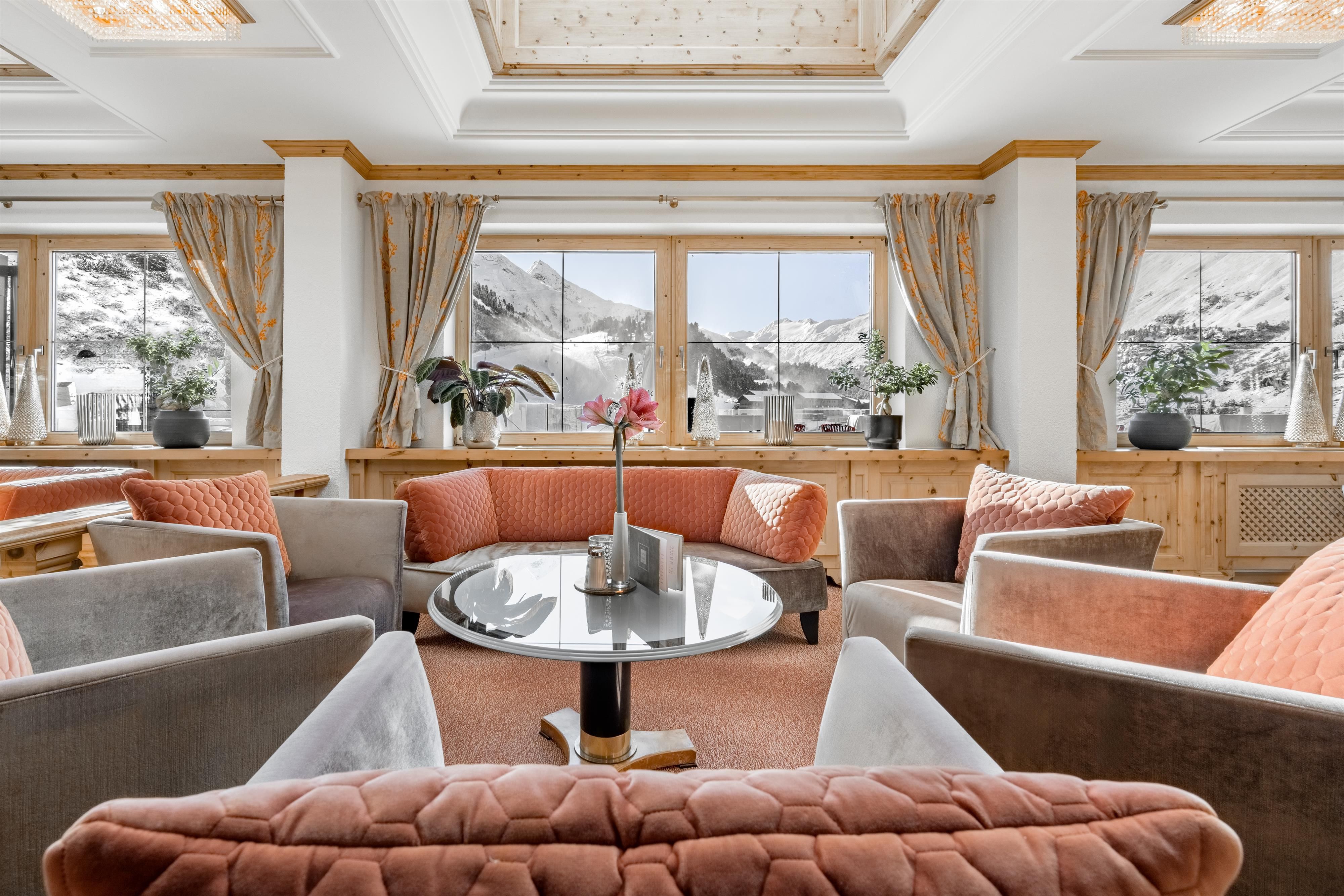 Ein eleganter Wohnbereich mit gemütlichen Sofas und einem zentralen Tisch. Große Fenster lassen viel Licht herein und bieten einen Blick auf die Berge.