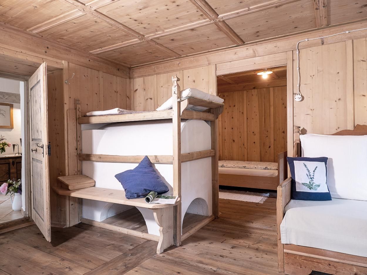 Originale Bauernstube mit Kachelofen, mit Holztäfelung und Holzfußboden, Durchsicht rechts auf das anschließende Schlafzimmer mit zwei getrennten Betten, links in den Eingangsbereich mit Holztruhe und Spiegel