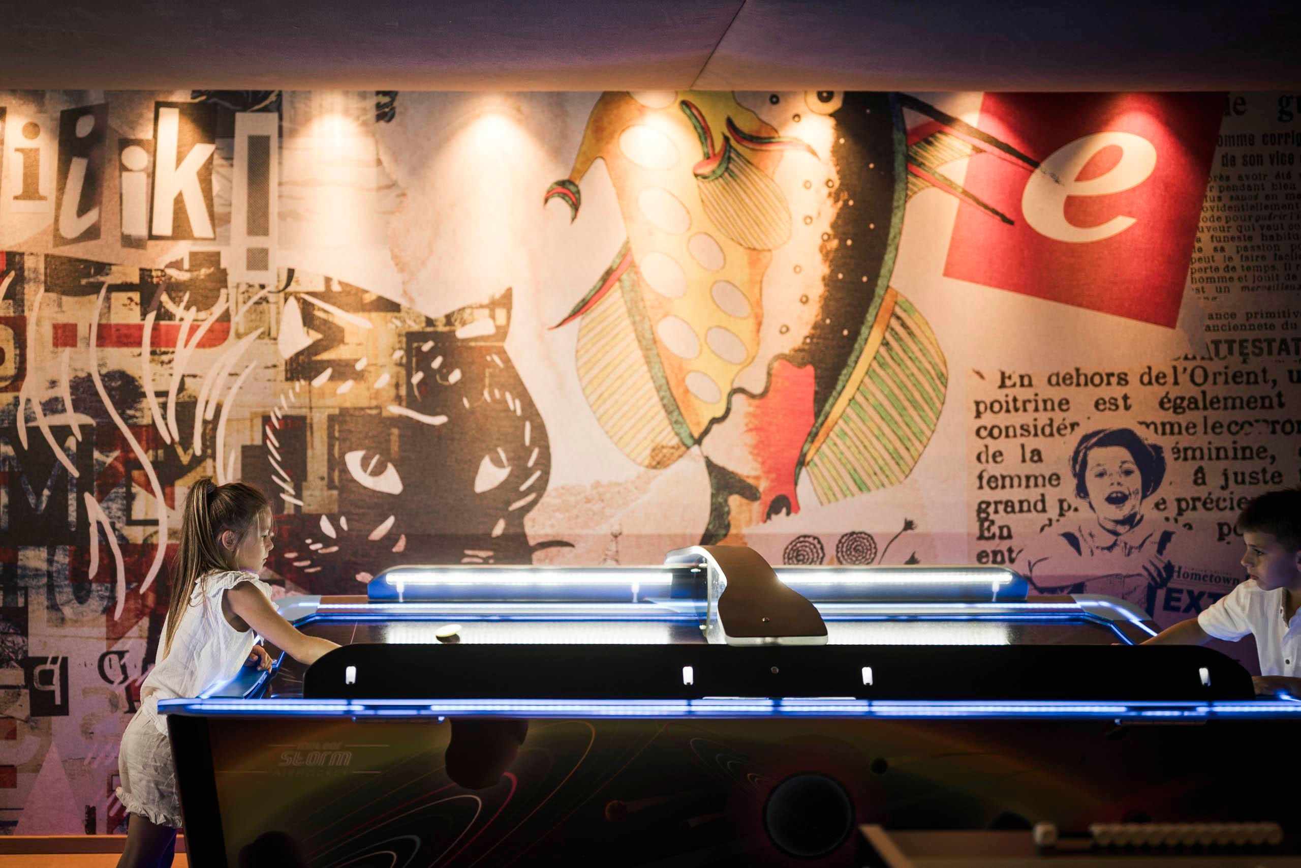 Zwei Kinder spielen an einem modernen Airhockey-Tisch. Im Hintergrund ist eine bunte Wand mit verschiedenen Grafiken und Texten zu sehen.