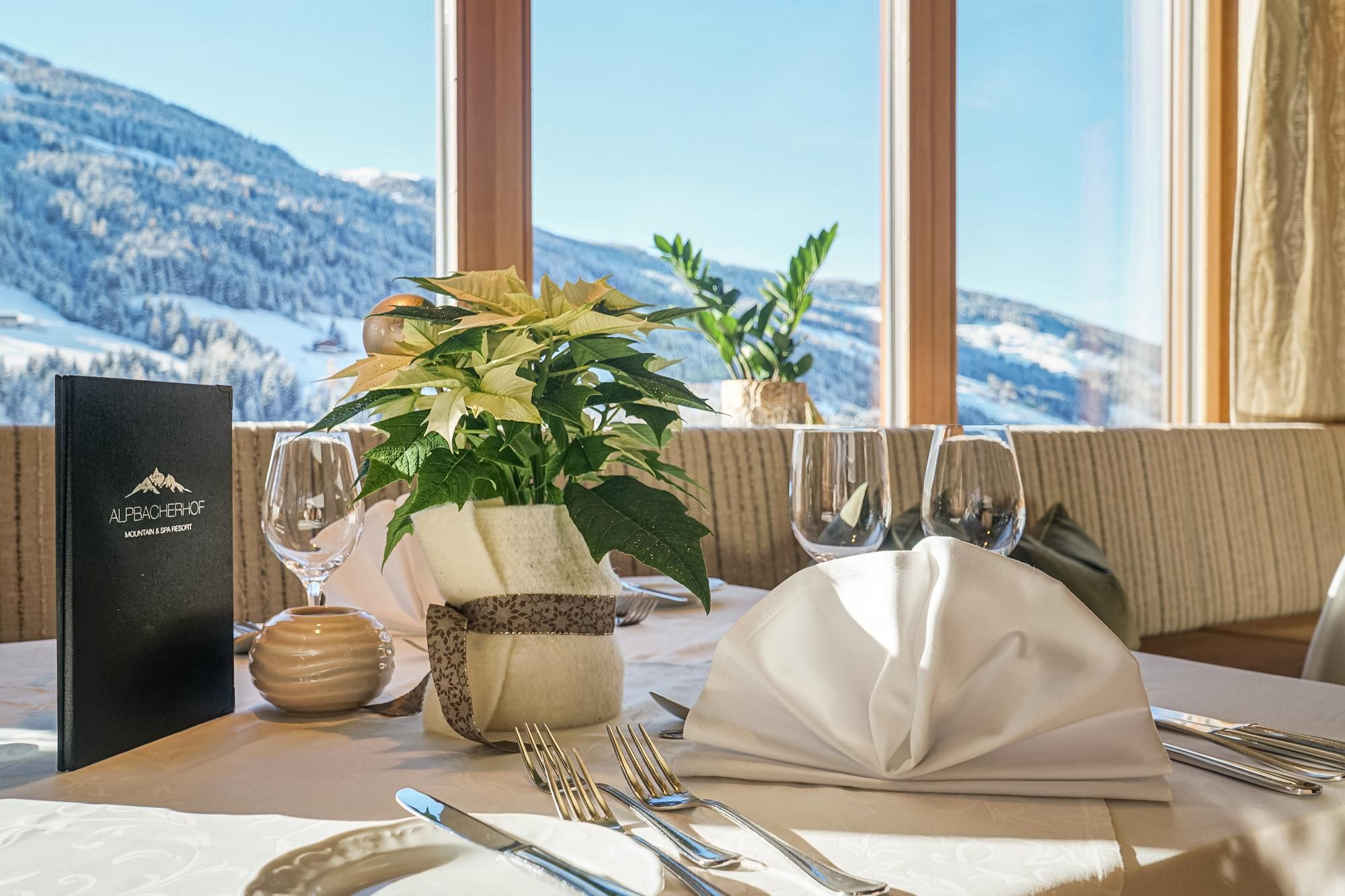 Ein elegant gedeckter Tisch im Restaurant mit Blick auf schneebedeckte Berge. Auf dem Tisch sind Pflanzen, Gläser und Besteck arrangiert.