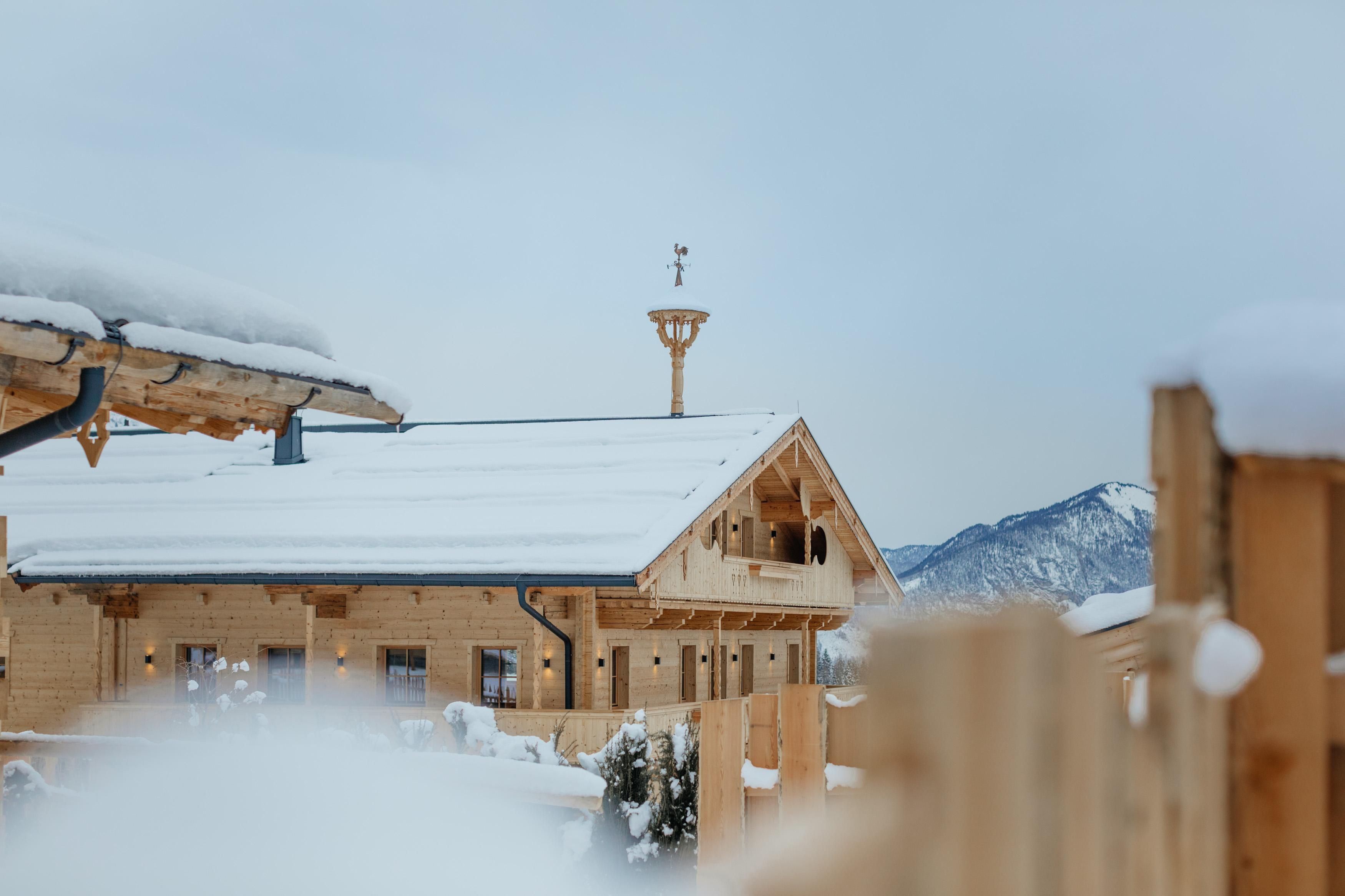 Ein gemütliches Holzhaus mit einem schneebedeckten Dach. Im Hintergrund sind schneebedeckte Berge zu sehen.