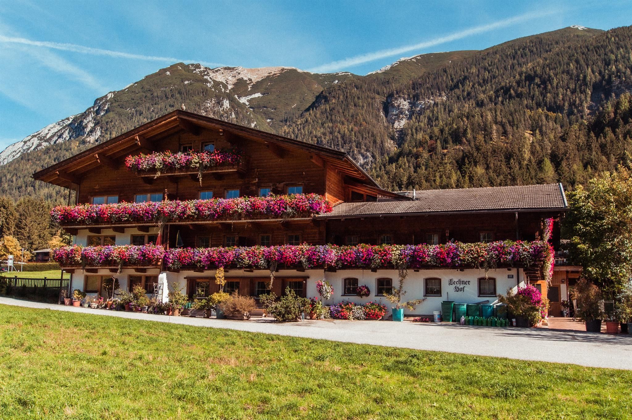 Ein traditionelles Alpenhaus mit blühenden Balkonkästen. Im Hintergrund sind schneebedeckte Berge und eine grüne Wiese zu sehen.
