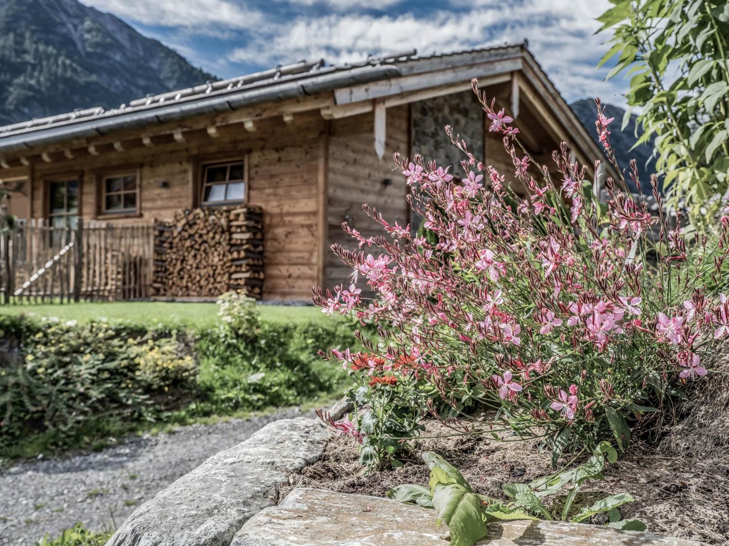 Ein traditionelles Chalet in einer malerischen Umgebung. Vor dem Haus blühen pinke Blumen und es ist eine grüne Wiese zu sehen.