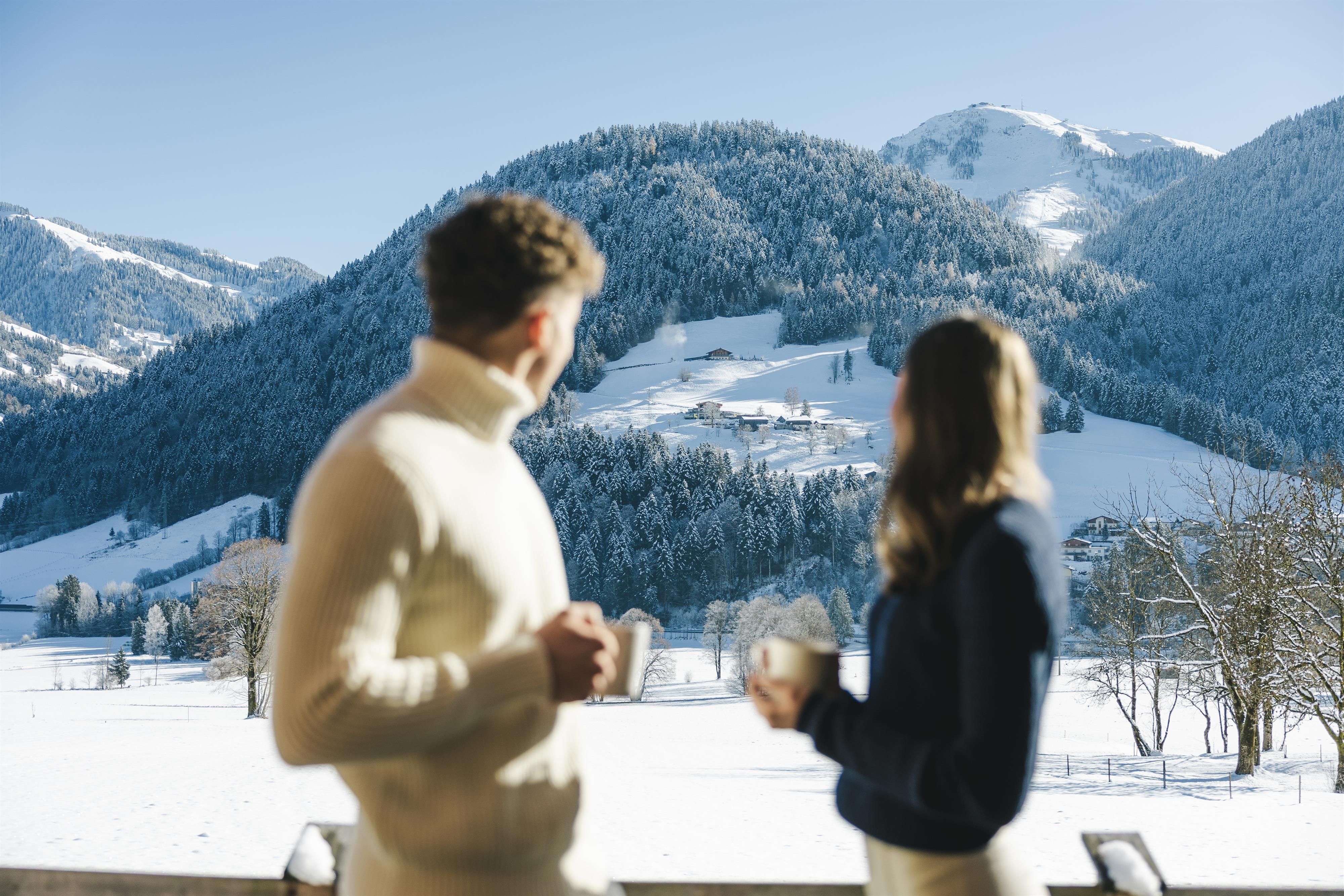 Ein Paar steht auf einem Balkon und genießt heiße Getränke. Im Hintergrund erstreckt sich eine verschneite Berglandschaft unter einem klaren blauen Himmel.