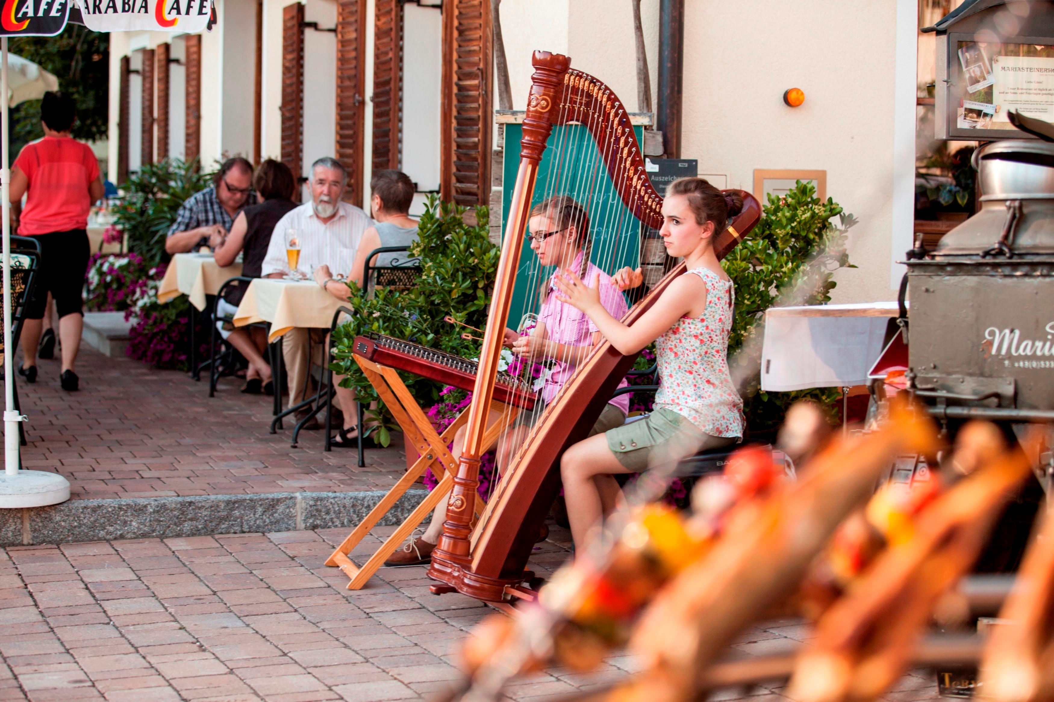 Eine junge Frau spielt Harfe in einem belebten Café. Gäste sitzen an Tischen und genießen die Atmosphäre im Freien.