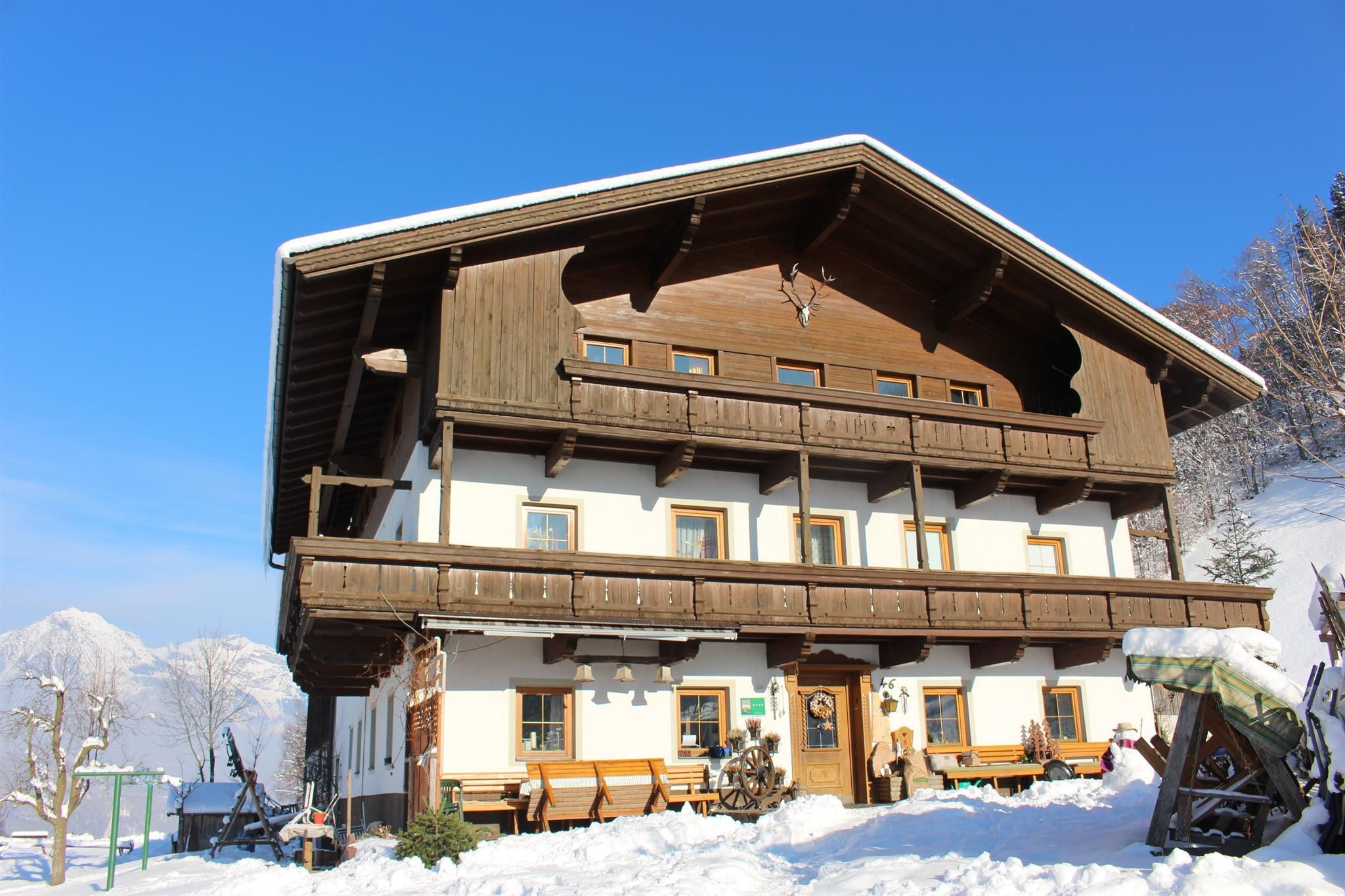Ein traditionelles Holzhaus im Schnee mit mehreren Fenstern und Balkonen. Der klare blaue Himmel rundet die winterliche Landschaft ab.
