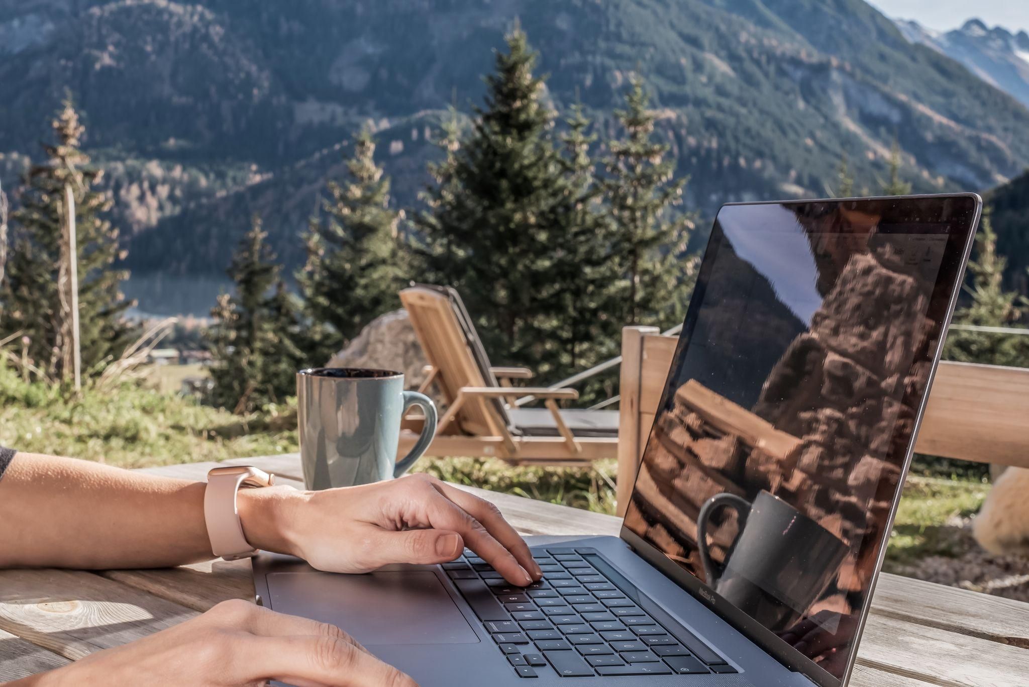 Eine Person arbeitet an einem Laptop im Freien, umgeben von schönen Bergen und Bäumen. Neben dem Laptop steht eine Tasse Kaffee.