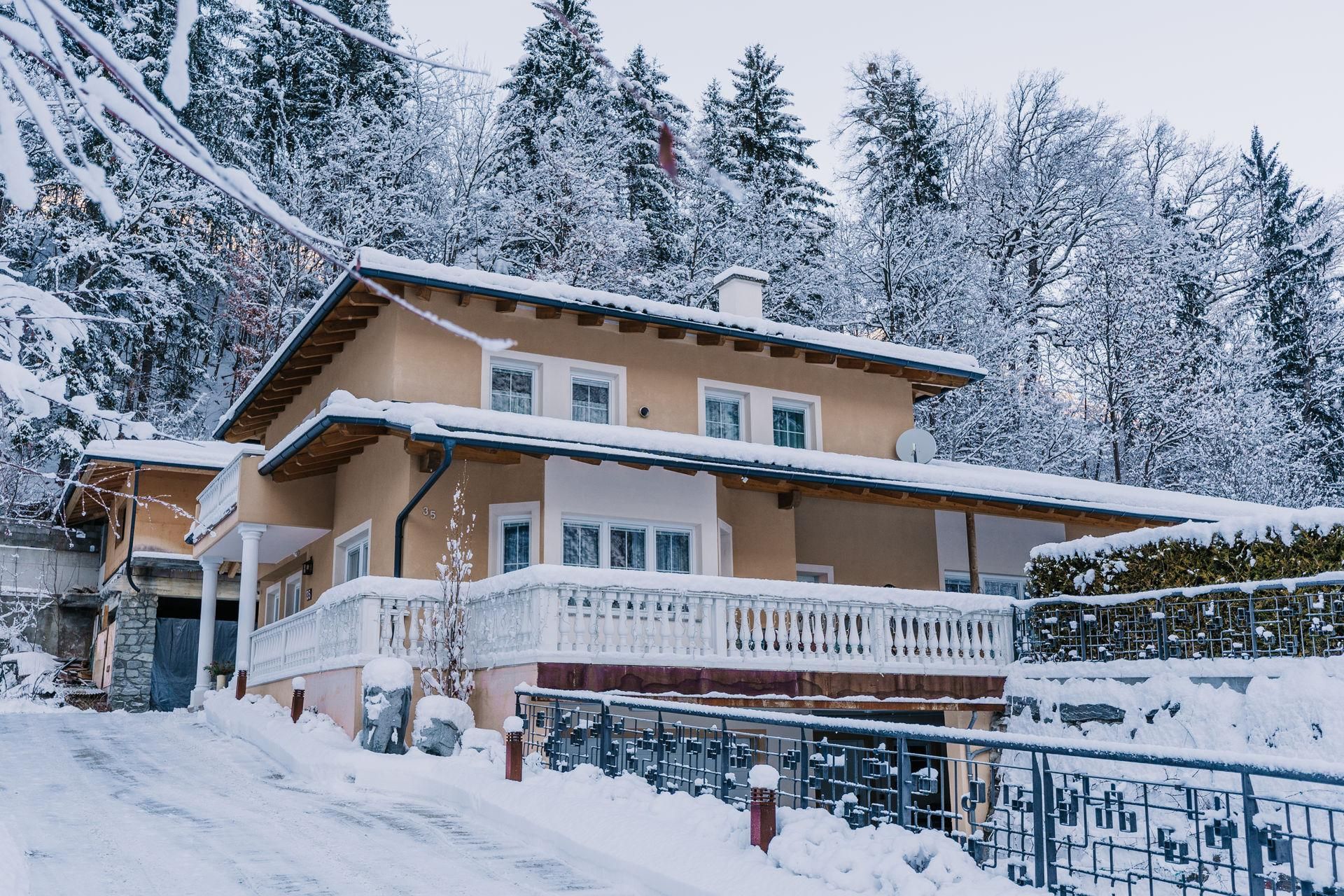 Unser schönes Haus im Winter mit schneebedecktem Dach und umgebenden Bäumen. Die Straße vor dem Haus ist ebenfalls mit Schnee bedeckt.