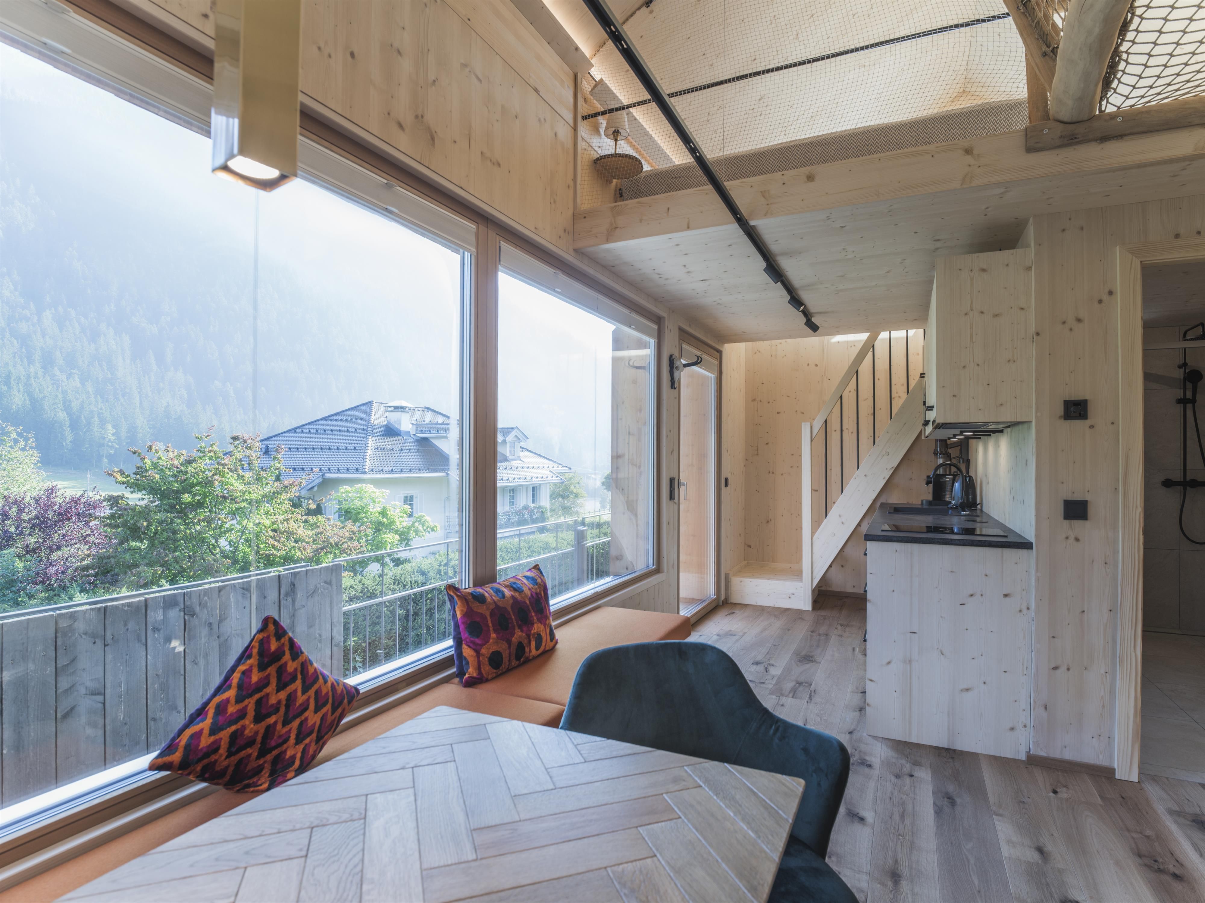 Ein modernes, helles Innendesign mit großen Fenstern und Holzverkleidungen. Der Raum zeigt eine gemütliche Sitzecke und eine Treppe zu einem oberen Bereich.