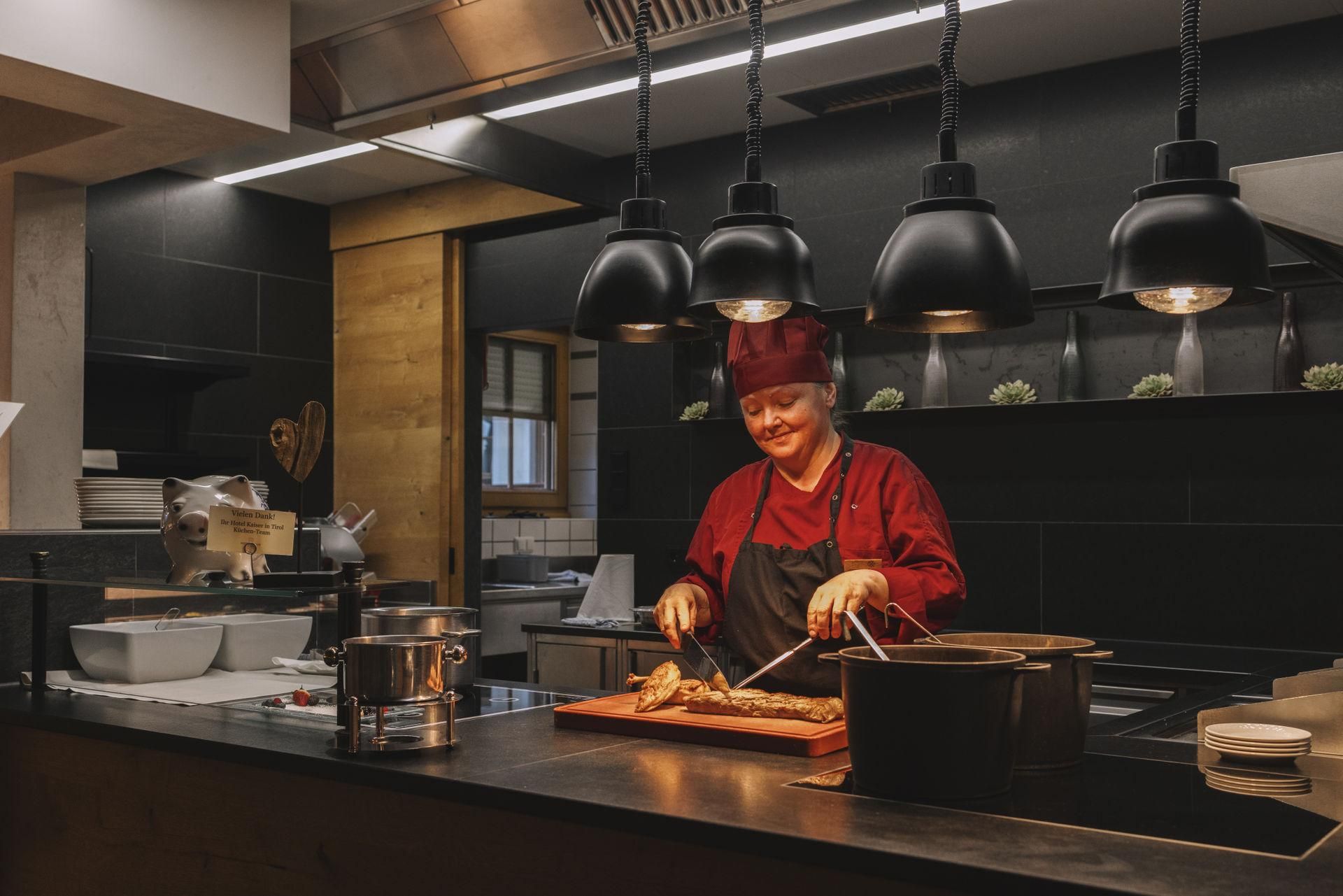 Ein Koch in einer modernen Küche bereitet Speisen zu. Er trägt eine rote Chefhut und lächelt, während er an einem Tisch arbeitet.