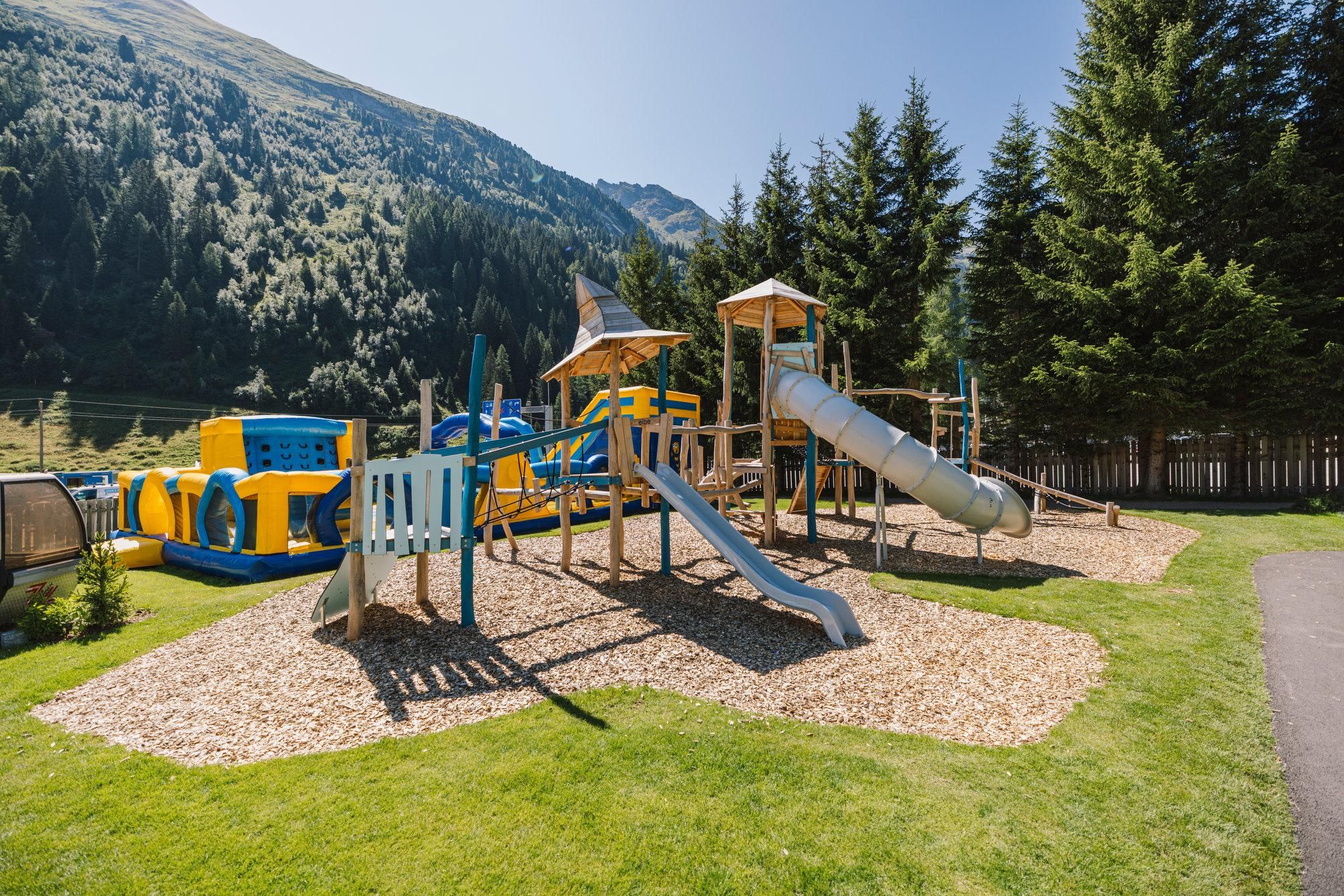 Ein Spielplatz mit Rutschen und Klettergerüsten, umgeben von Grünflächen und Bäumen. Im Hintergrund sind Berge zu sehen.
