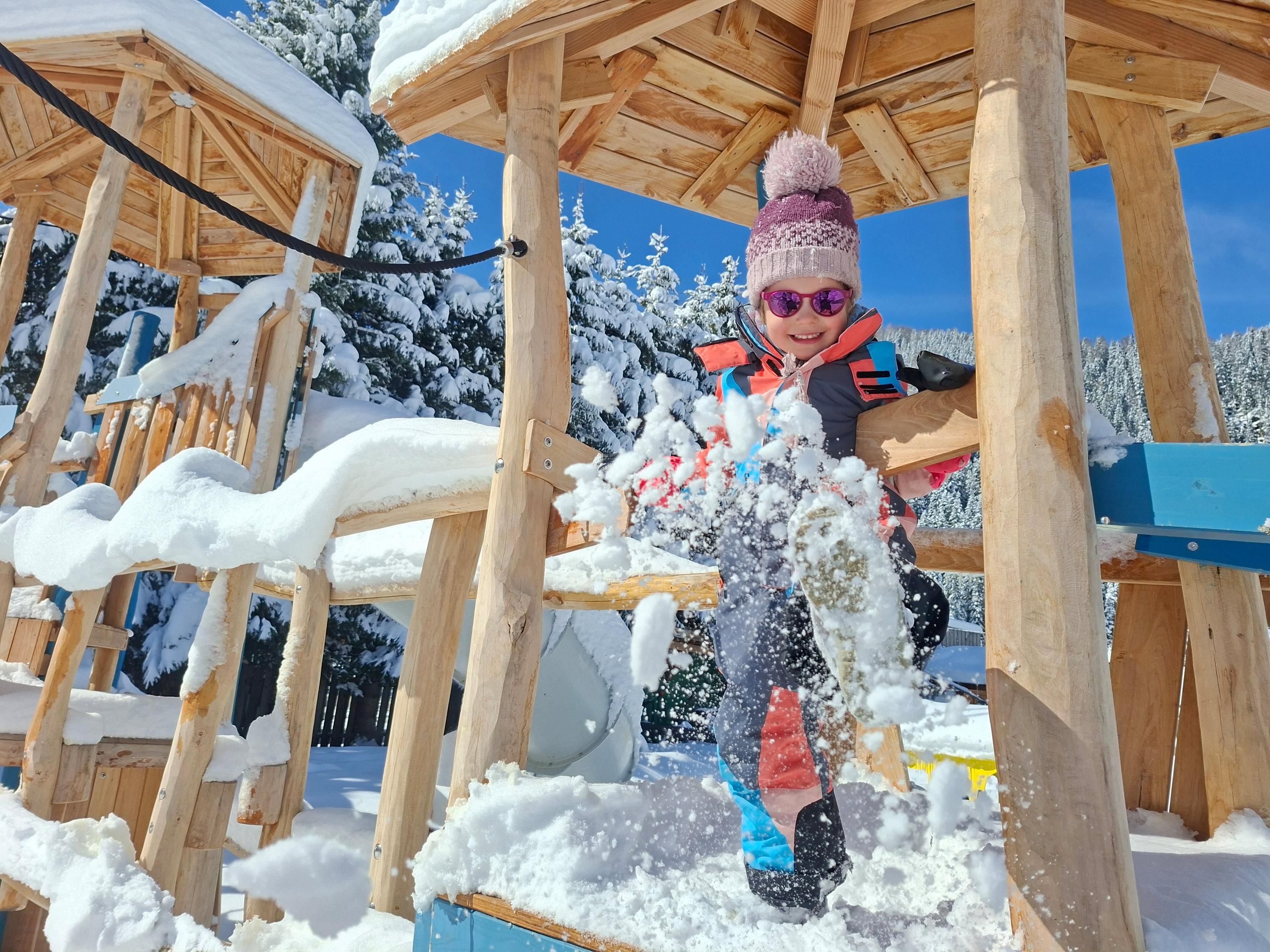 Ein Kind spielt fröhlich im Schnee auf einem Spielplatz. Die Umgebung ist winterlich mit viel Schnee und blauen Himmel.