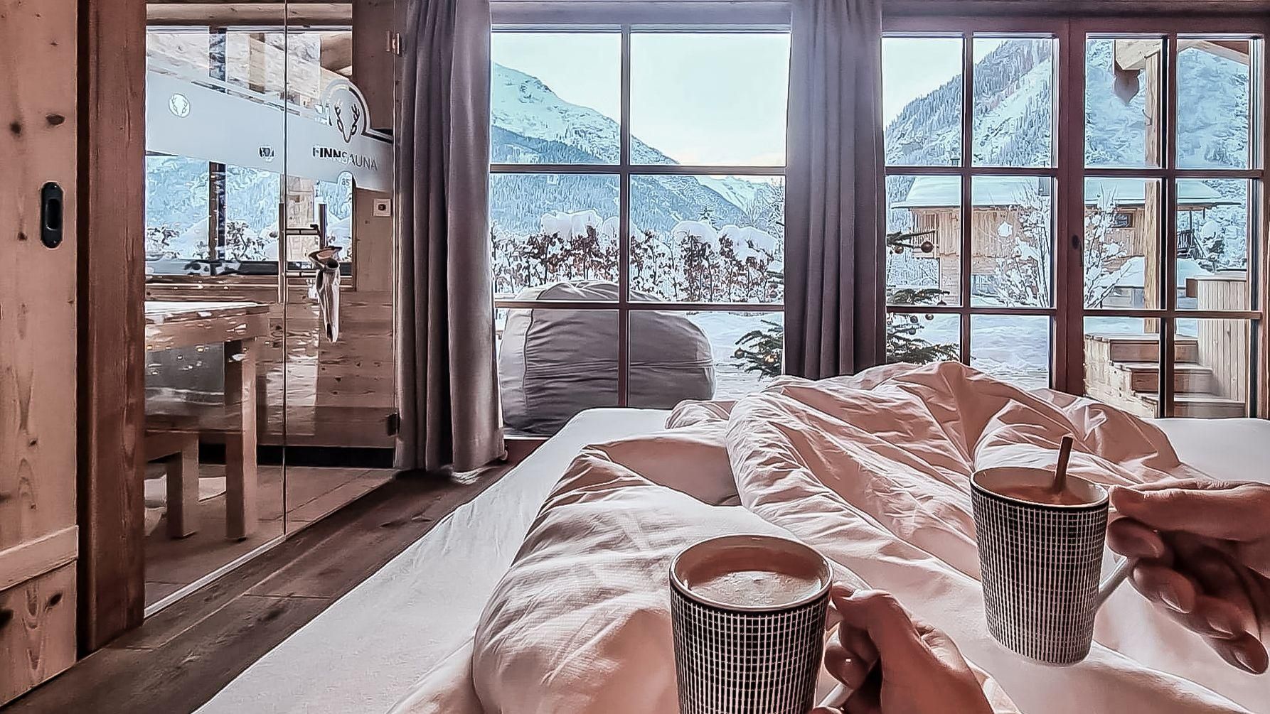 Ein gemütliches Schlafzimmer mit einem Blick auf die Berge. Zwei Tassen heißen Getränks werden inmitten frischer Bettwäsche gehalten.