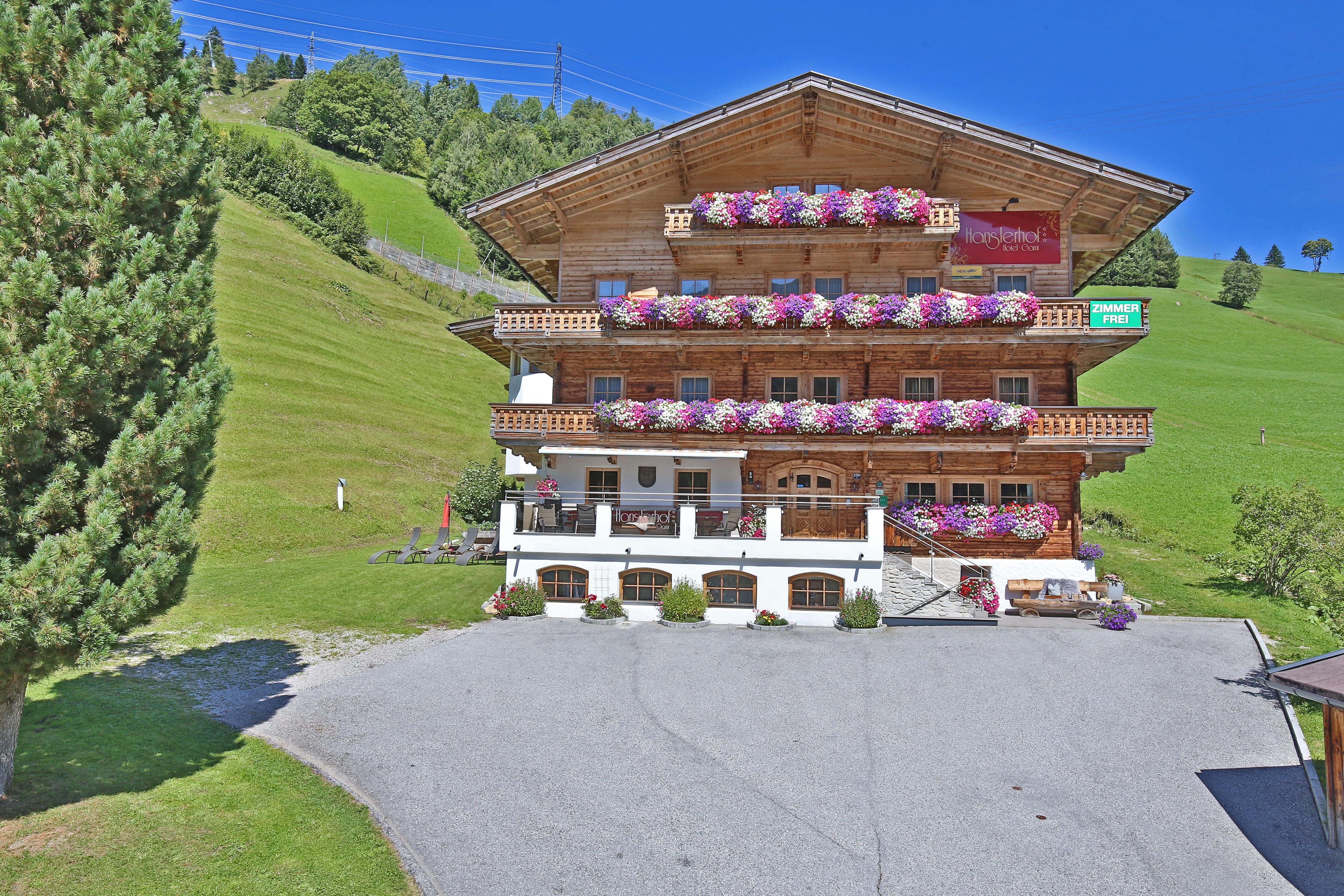 Ein charmantes Holzhaus mit blühenden Balkonpflanzen. Umgeben von grünen Wiesen und einem klaren blauen Himmel.