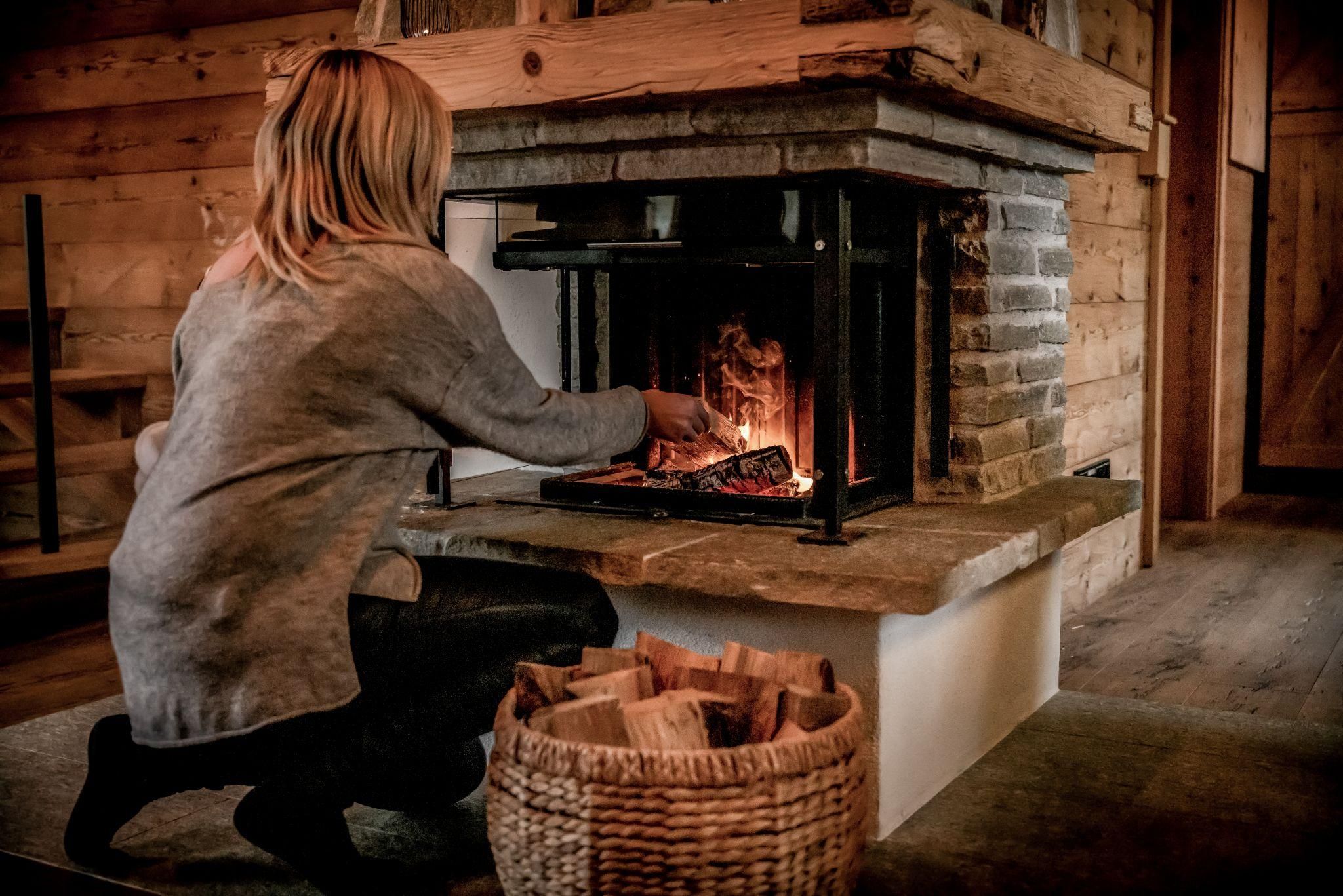 Eine Person sitzt vor einem Kamin und füttert das Feuer mit Holz. Im Hintergrund sind Holzverkleidungen und ein Korb mit Holzscheiten zu sehen.