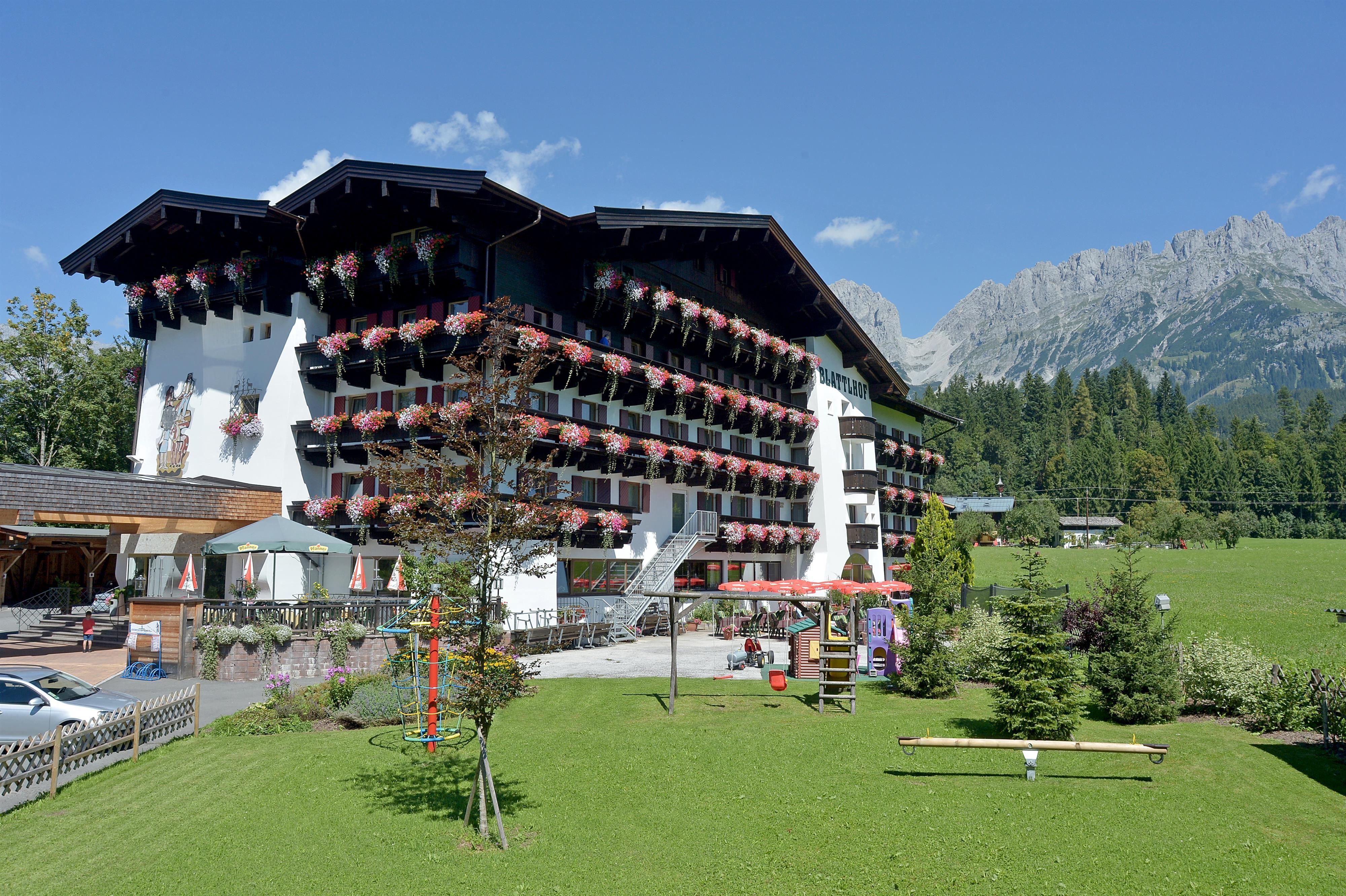 Ein malerisches Hotelgebäude mit Balkonen, die mit Blumen geschmückt sind. Im Hintergrund sind grüne Wiesen und beeindruckende Berge zu sehen.