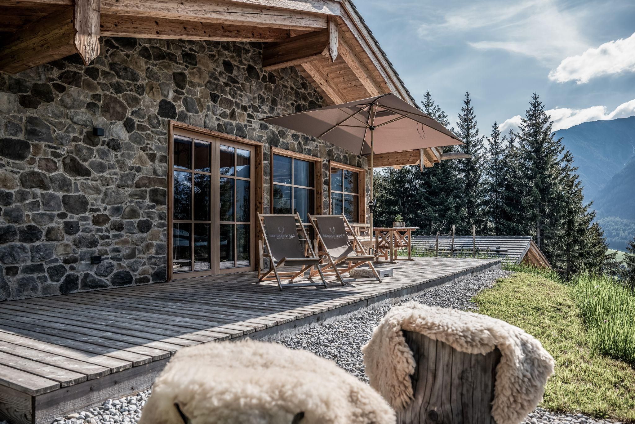 Ein modernes Chalet aus Stein mit einer Holzterrasse. Im Vordergrund stehen Liegestühle und ein Sonnenschirm, umgeben von einer malerischen Berglandschaft.