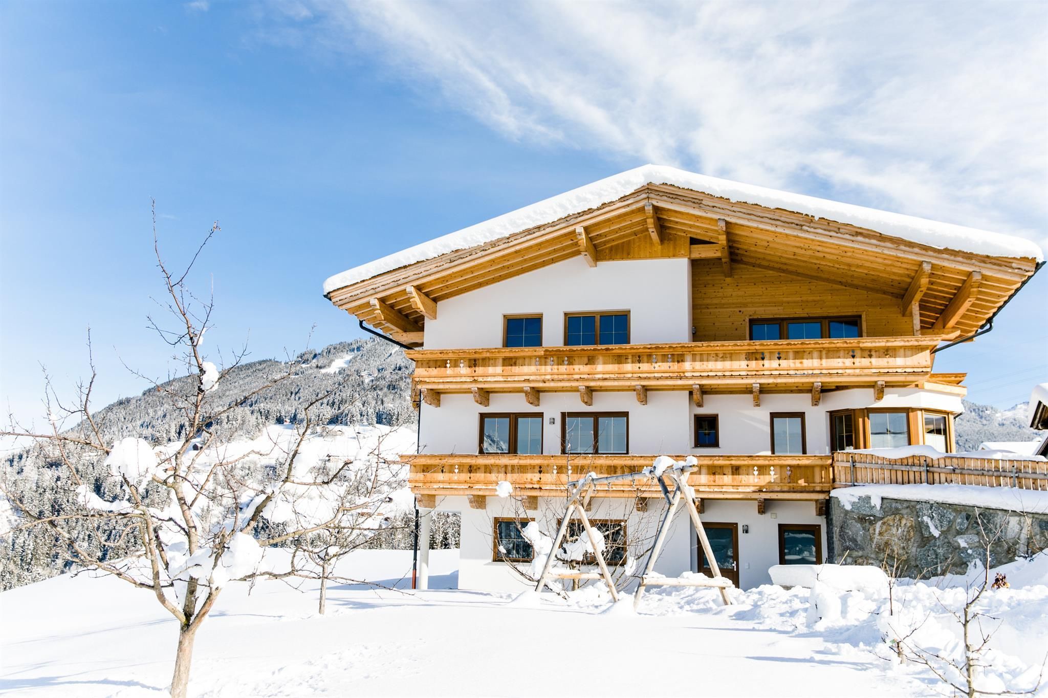 Ein schönes Haus im Schnee mit einem traditionellen Holzbalcony. Im Hintergrund sind schneebedeckte Berge und ein klarer blauer Himmel zu sehen.