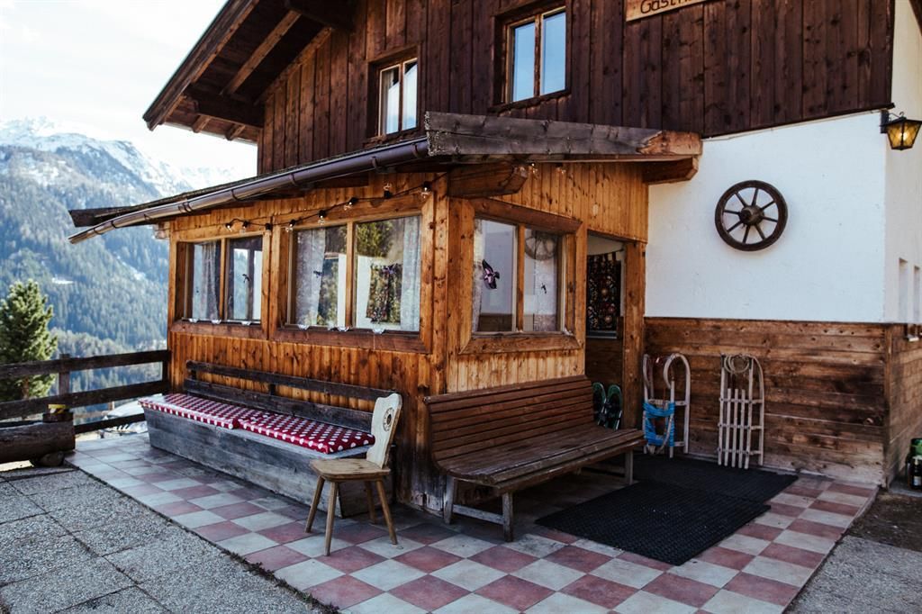 Eine gemütliche Berghütte aus Holz mit einem überdachten Bereich und Fenstern. Im Hintergrund sind die Berge sichtbar, und der Boden ist mit bunten Fliesen gestaltet.
