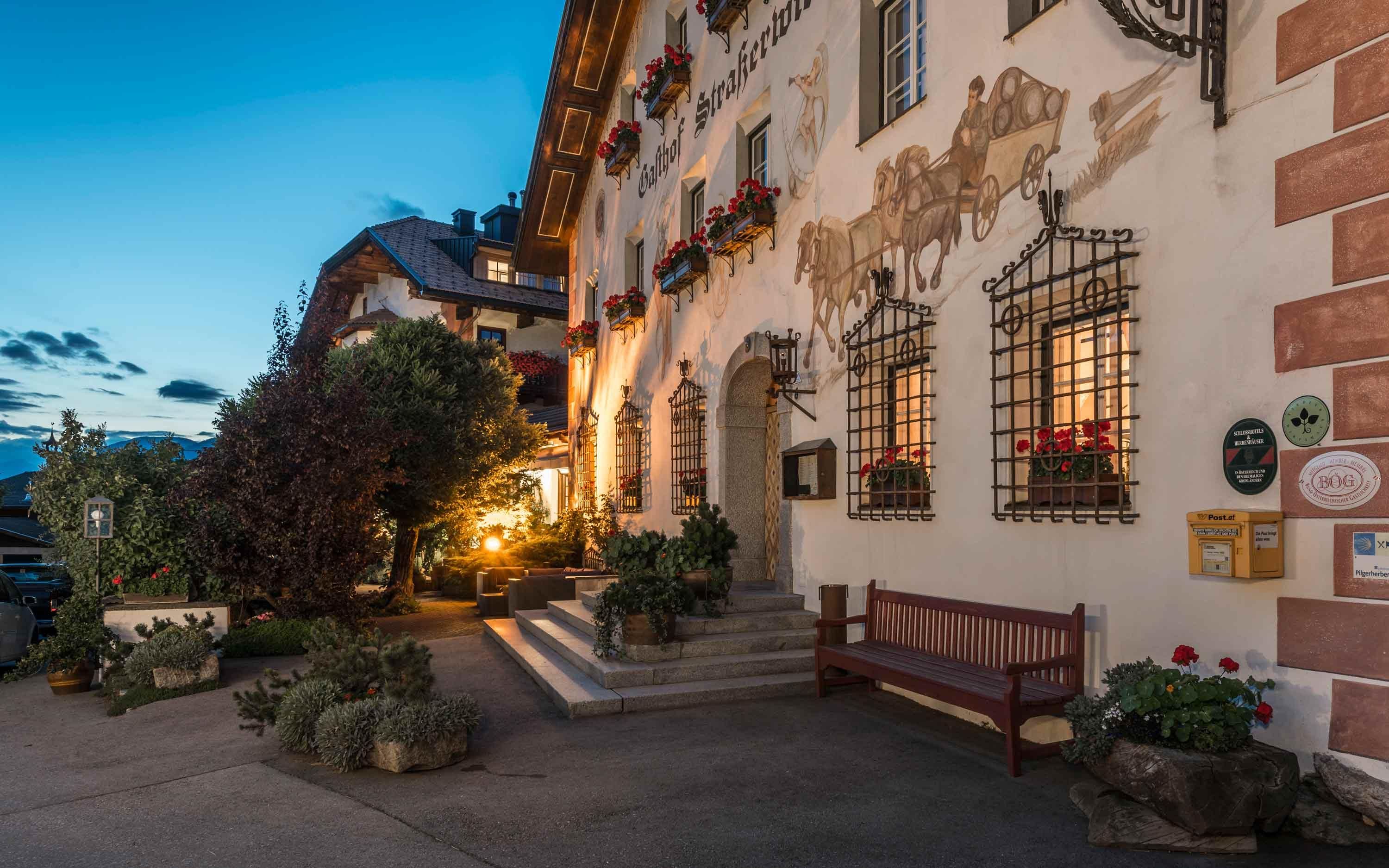 Der Strasserwirt, ein traditioneller Hotel am Abend. Die Fassade ist bemalt mit einer Pferdewagenszene. Rote Blumen in Kästen hängen an Fenstern mit Eisenstäben. Eine rote Bank steht am Eingang, daneben ein gelber Postkasten. Warme Beleuchtung und viele Pflanzen schaffen eine einladende Atmosphäre. Im Hintergrund Berge und Bäume.