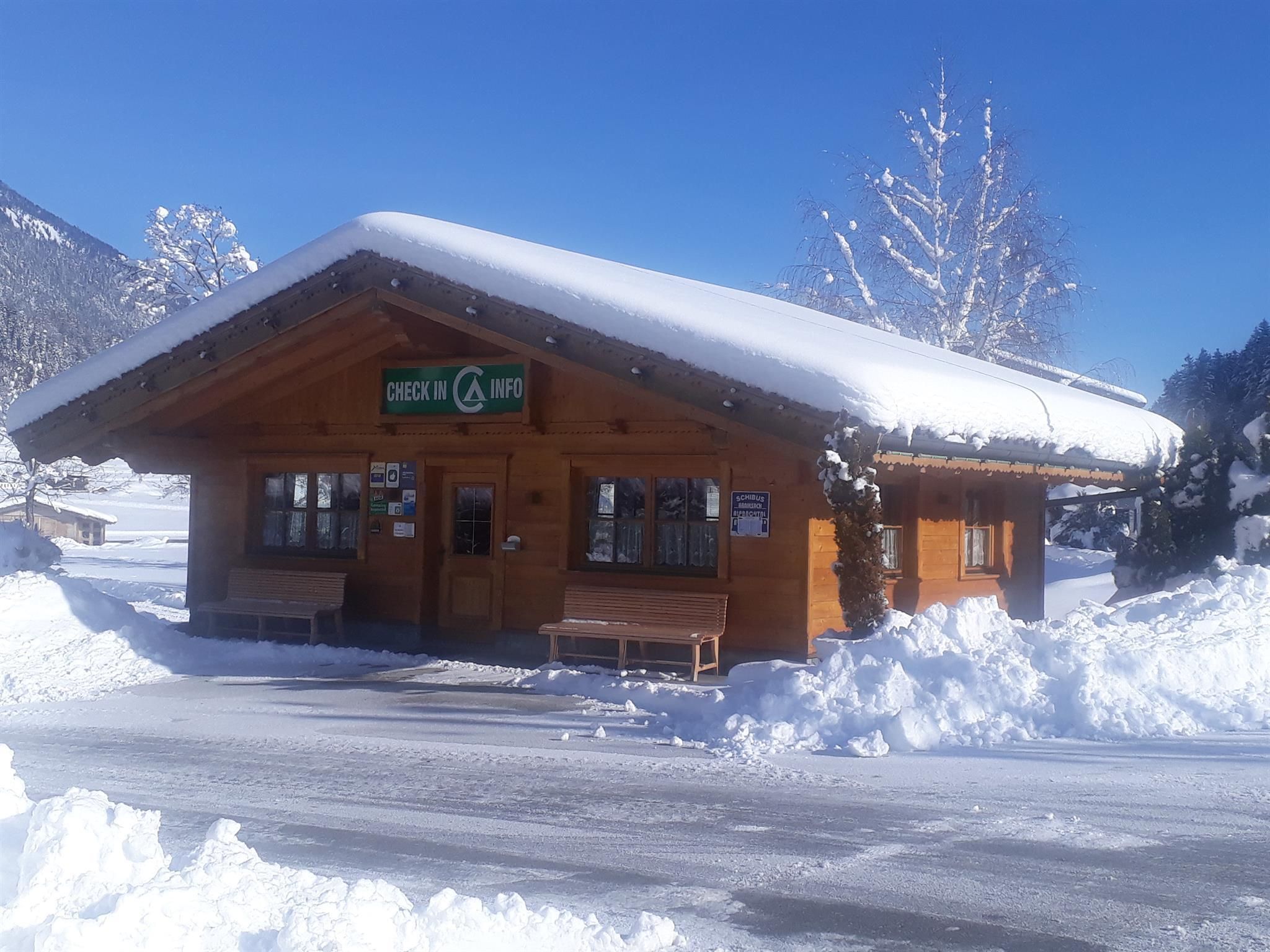 Ein charmantes Holzhaus im Schnee, umgeben von verschneiten Bäumen. Der Himmel ist klar und blau.