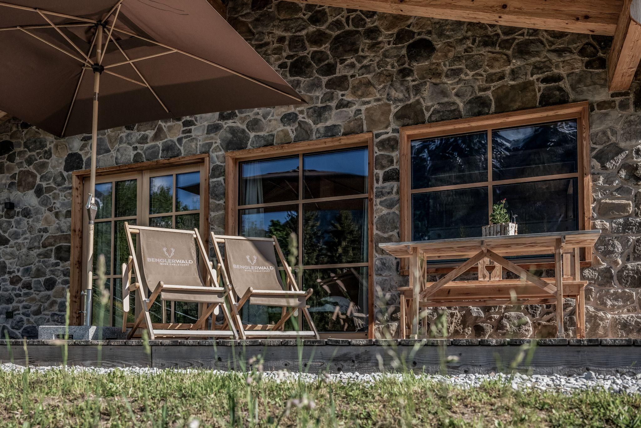 Ein gemütlicher Außenbereich mit Sonnenliegen und einem Holztisch. Im Hintergrund steht ein rustikales Stein Chalet mit großen Fenstern.