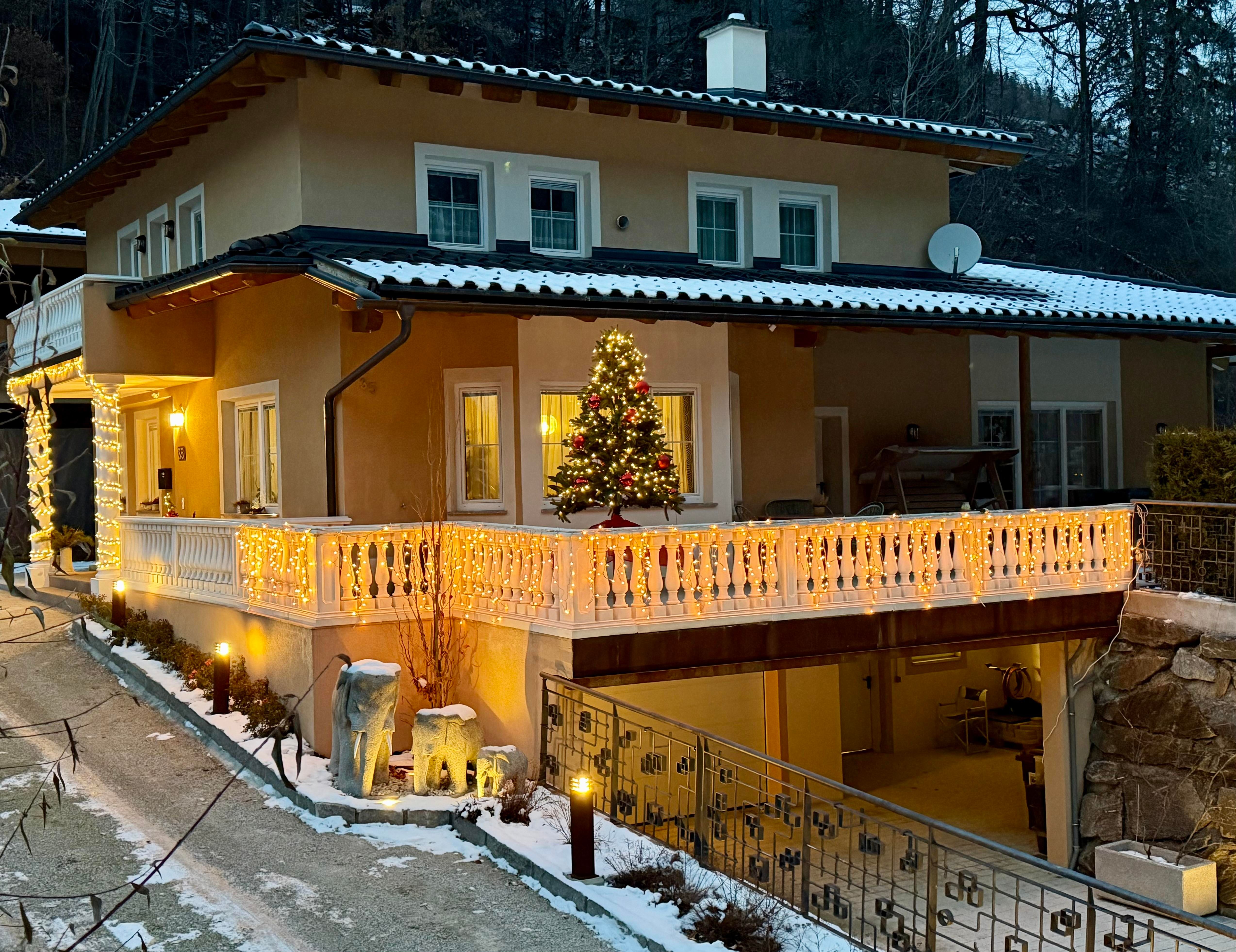 Ein schönes Haus mit Weihnachtsdekoration und einem beleuchteten Balkon. Schnee bedeckt den Boden und die Bäume im Hintergrund.