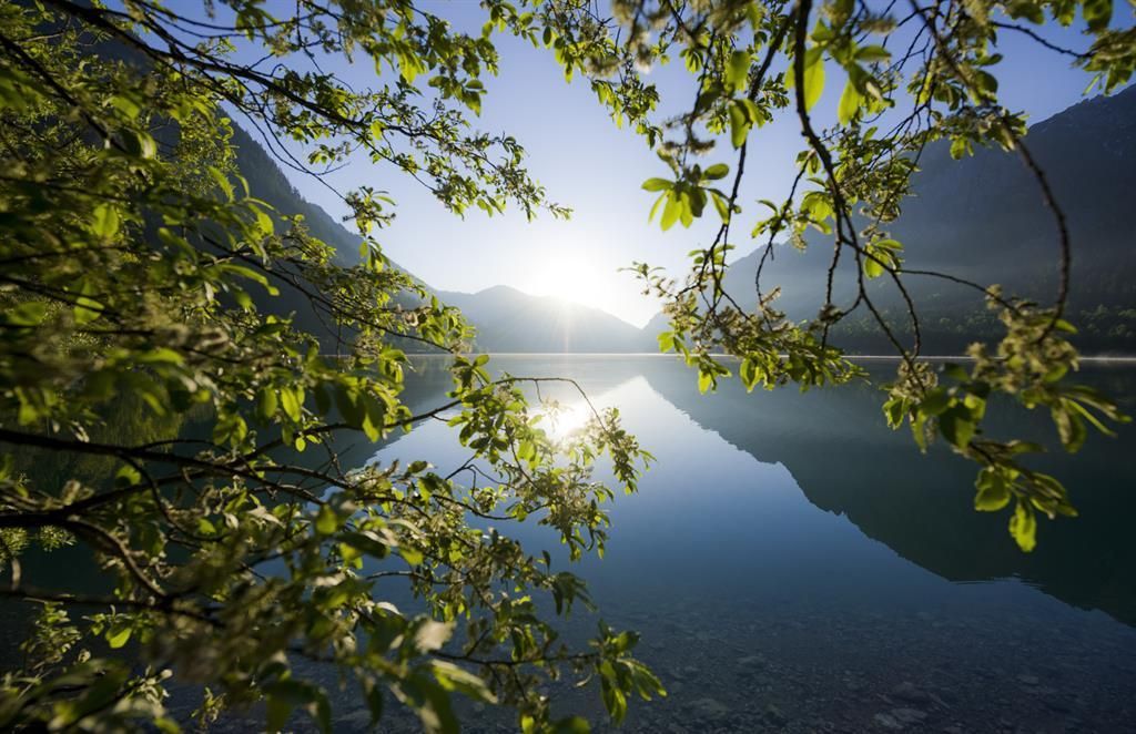 Eine ruhige Seenlandschaft, umgeben von Bergen und Bäumen. Die Sonne spiegelt sich im klaren Wasser.