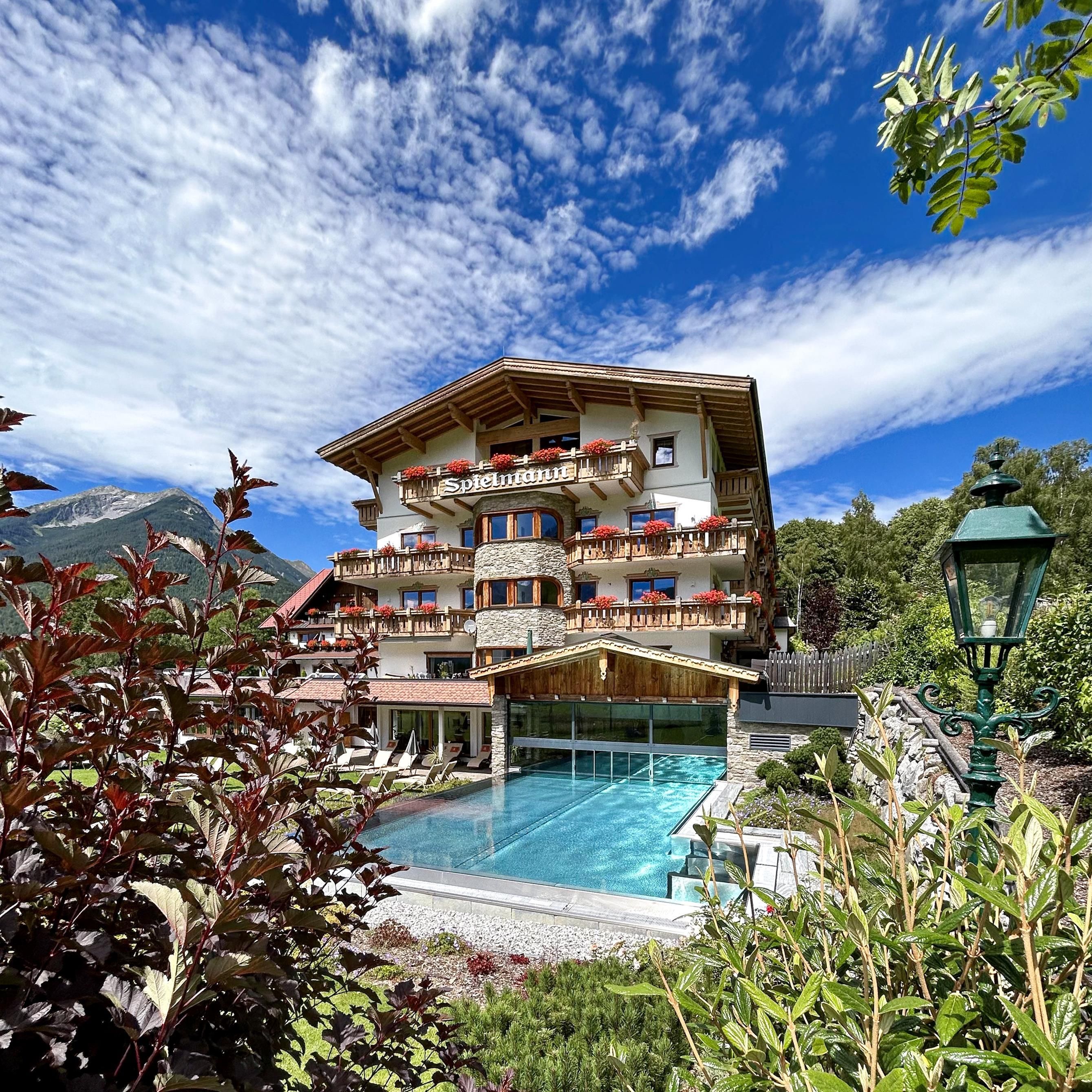 Ein schönes Hotel mit Balkonen und einem Pool im Vordergrund. Der Himmel ist klar mit einigen Wolken und die Umgebung ist grün und einladend.