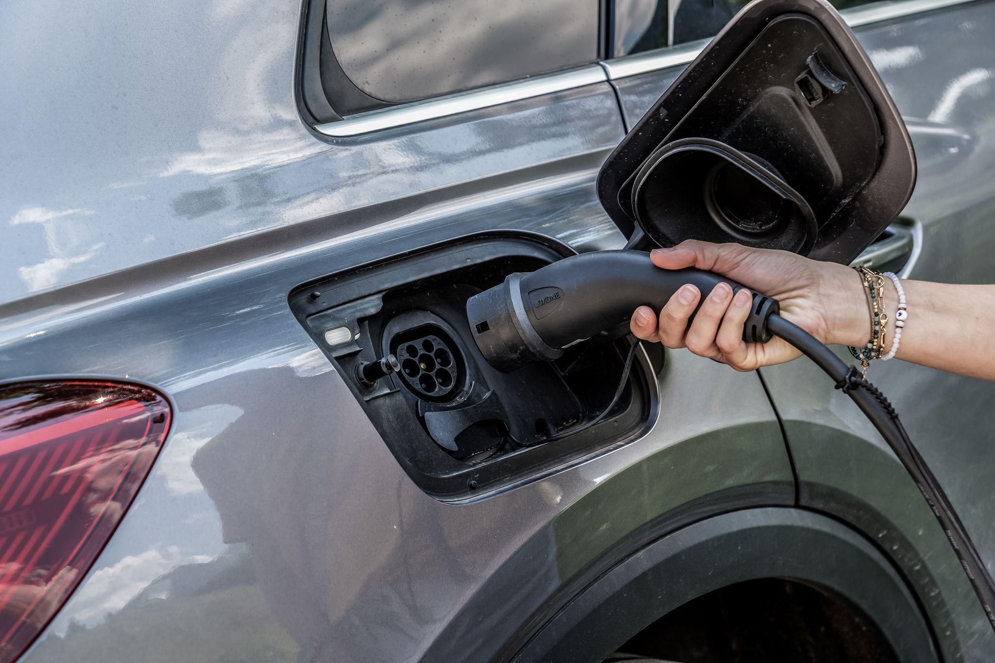 Eine Hand, die ein Ladekabel in die Ladebuchse eines grauen Elektrofahrzeugs steckt. Der Hintergrund zeigt das Design des Autokörpers.