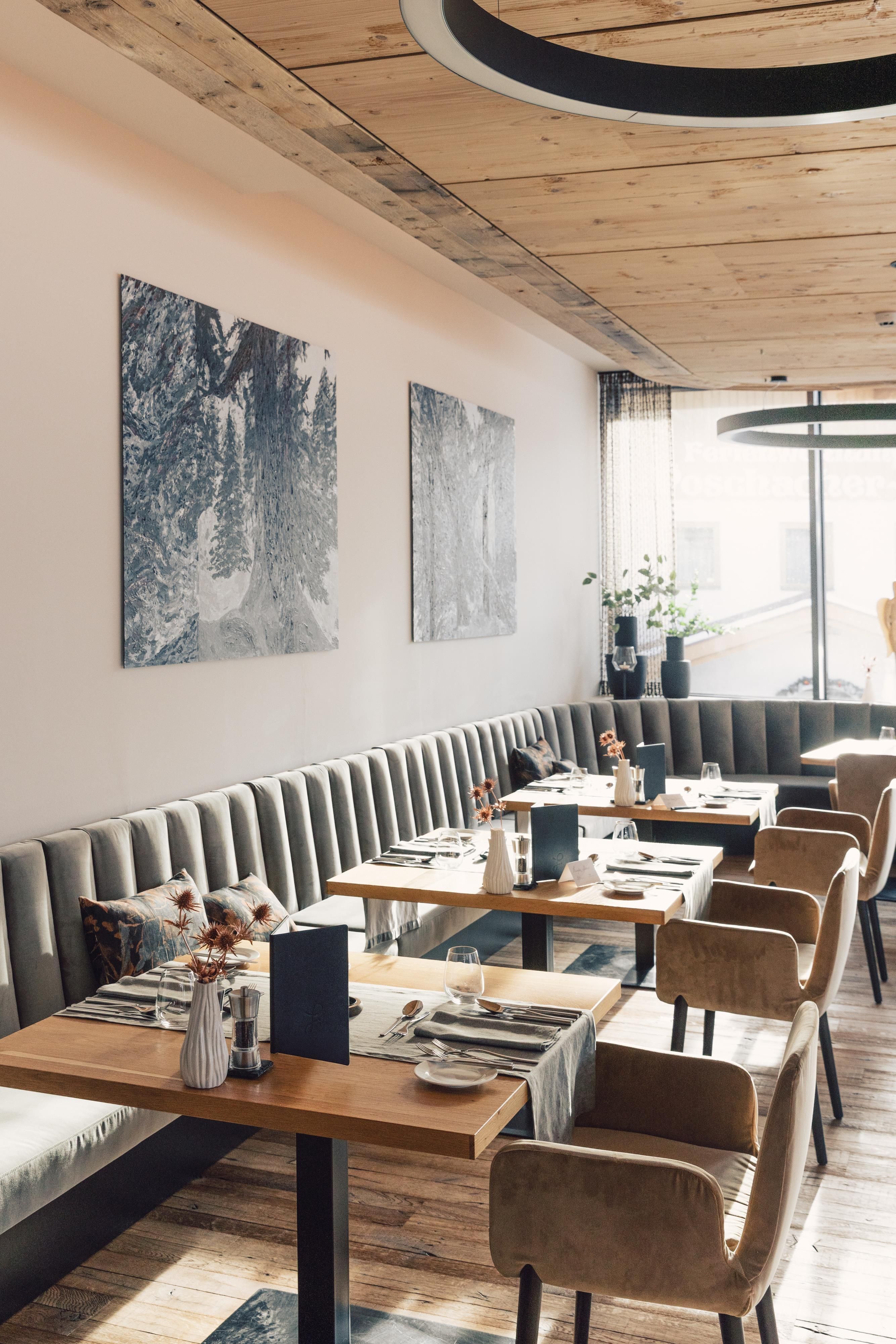 Ein modernes Restaurant mit eleganten Tischdekorationen und bequemen Sitzen. An den Wänden hängen kunstvolle Bilder, und große Fenster sorgen für viel Tageslicht.