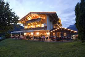 Ein gemütliches Haus im alpinen Stil, beleuchtet bei Dämmerung. Im Vordergrund gibt es eine Terrasse mit Sitzplätzen im Freien.