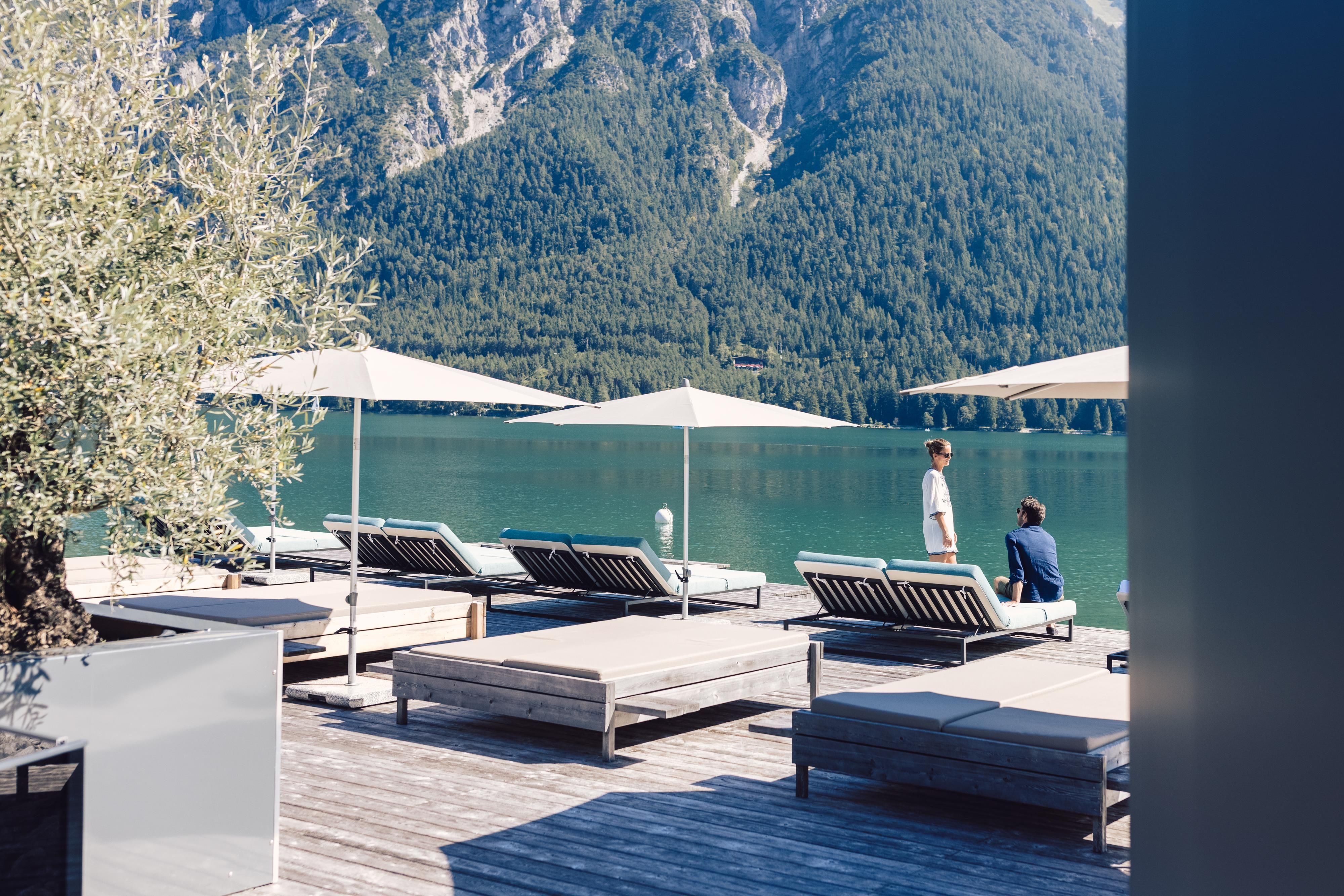 Ein ruhiger See mit Sonnenliegen und Sonnenschirmen. Im Hintergrund sind beeindruckende Berge zu sehen.