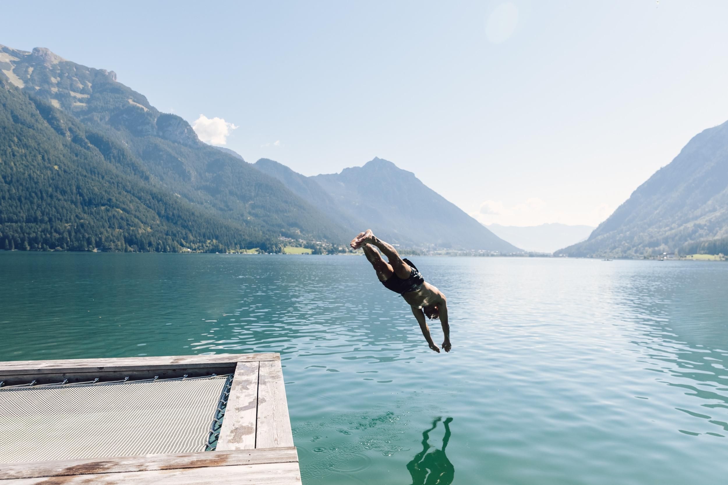Ein Mann springt von einem Steg in einen klaren, blauen See. Im Hintergrund sind majestätische Berge und ein strahlend blauer Himmel zu sehen.