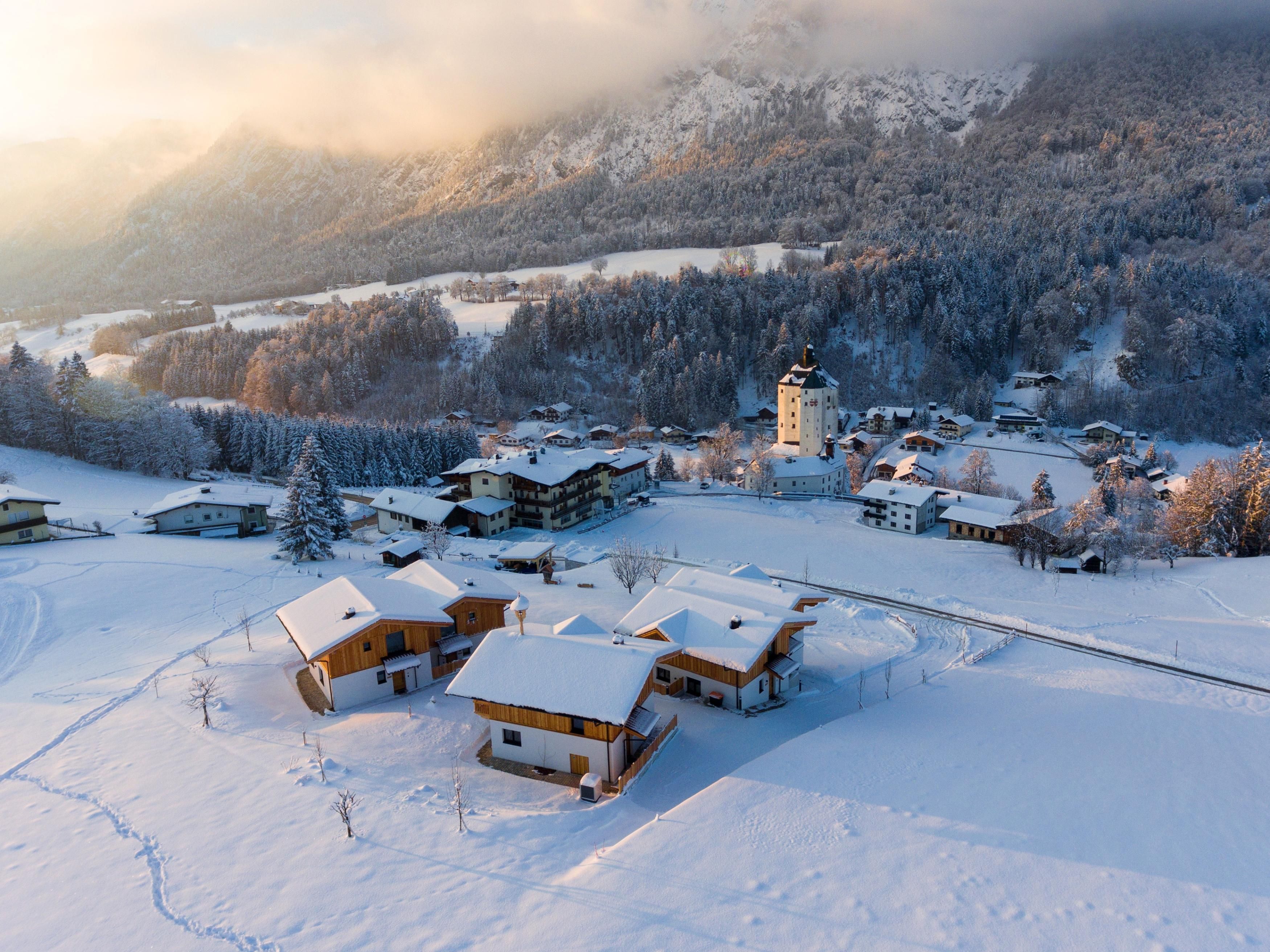 Eine verschneite Landschaft mit traditionellen Hütten und vielen Bäumen. Im Hintergrund sind Berge und eine klare Winteratmosphäre zu sehen.