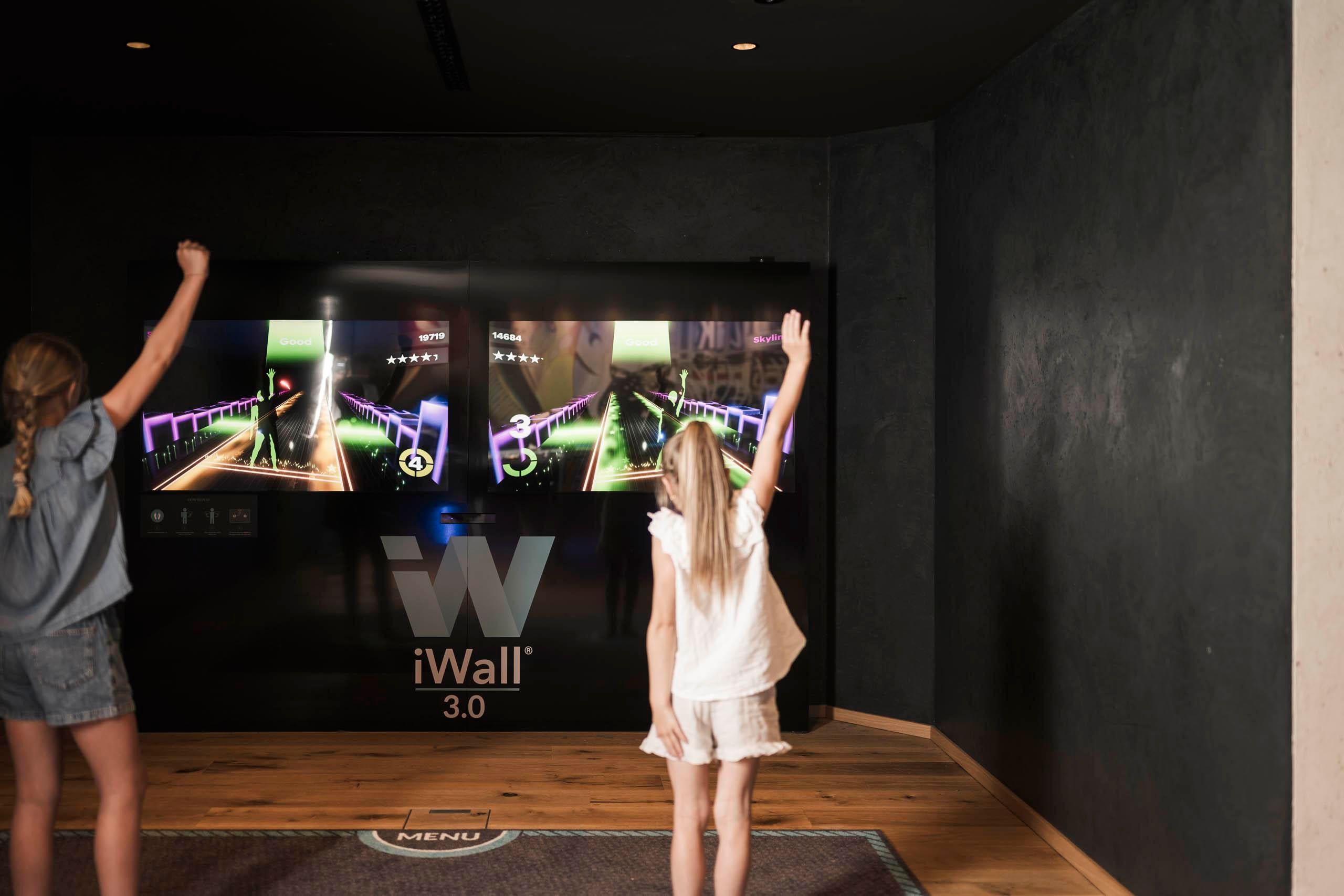 Zwei Mädchen spielen ein interaktives Spiel an einem großen Bildschirm. Sie stehen vor der Wand mit bunten Grafiken und haben ihre Arme gehoben.