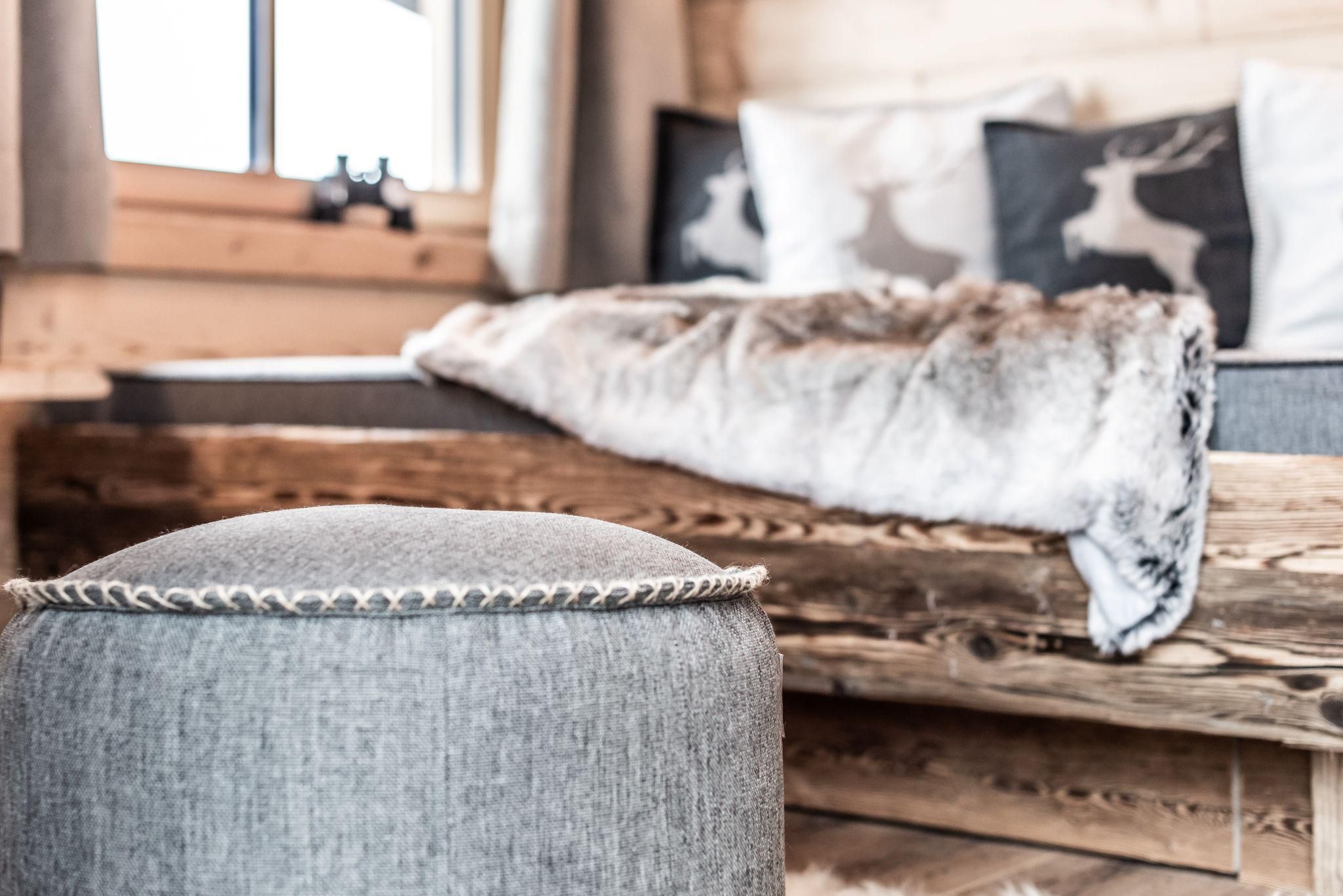 Ein gemütliches Zimmer mit einem Holzbett und dekorativen Kissen. Ein grauer Hocker steht im Vordergrund, während eine Decke auf dem Bett liegt.
