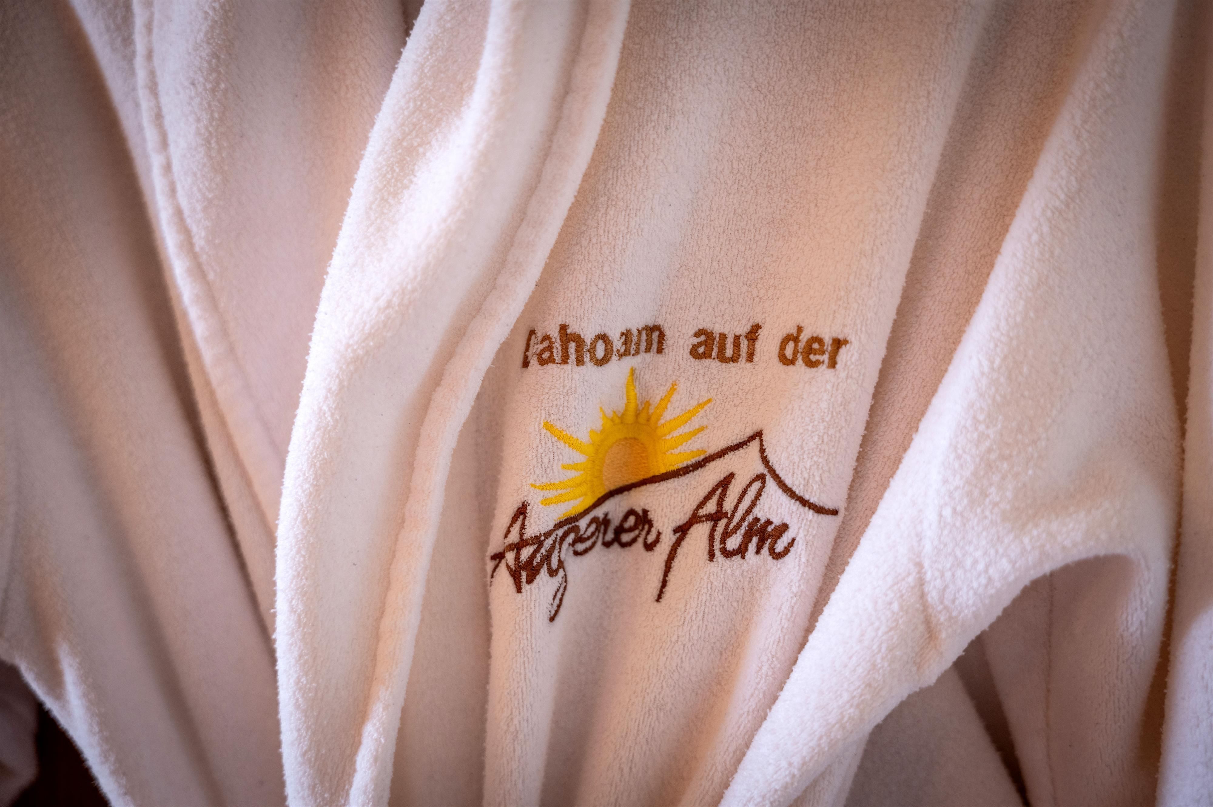Ein weicher Bademantel mit dem Aufdruck "Zuhause auf der Angerer Alm" und einer sonnigen Grafik. Der Bademantel vermittelt ein Gefühl von Gemütlichkeit und Entspannung.