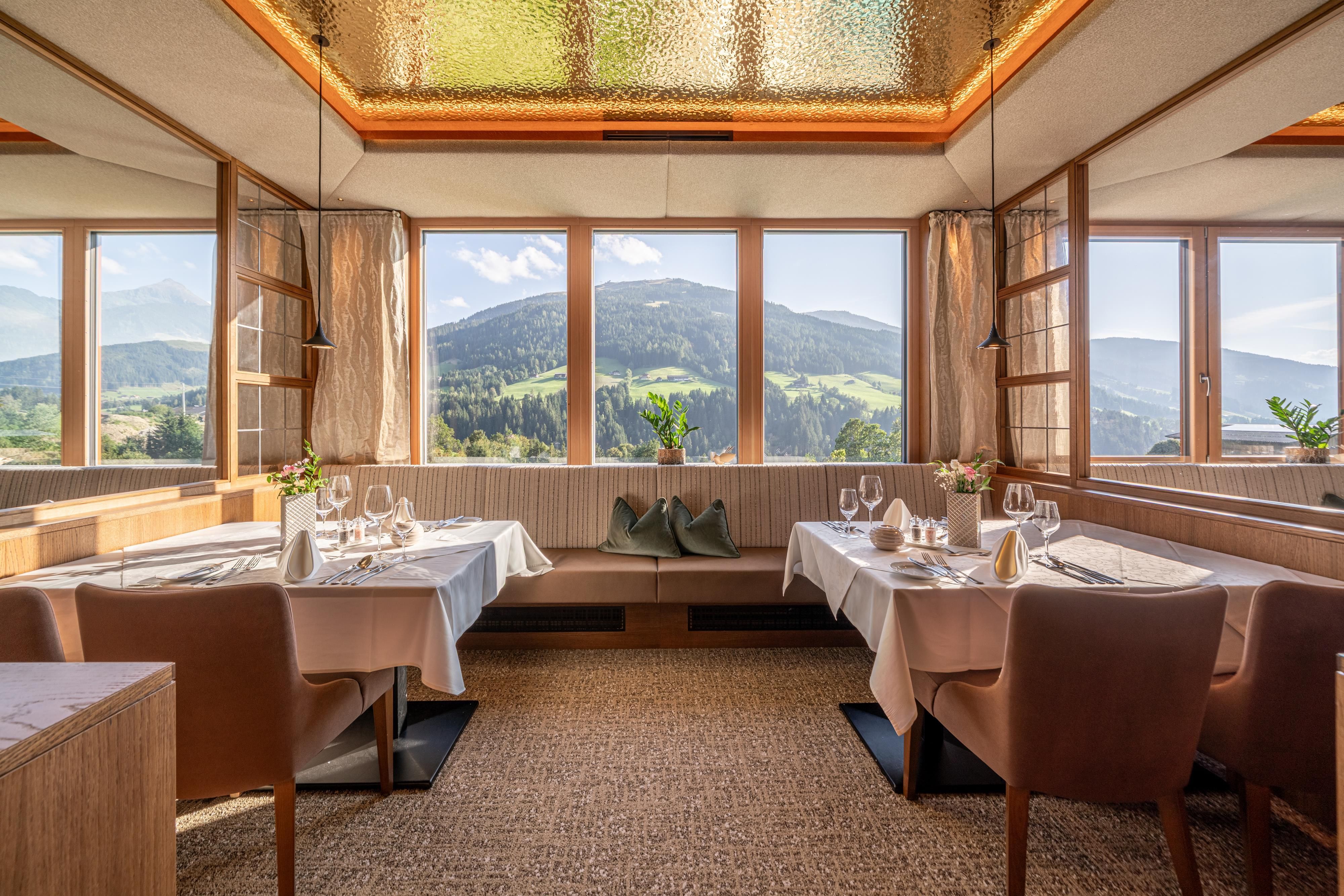 Ein elegantes Restaurant mit Tischen, die weiß eingedeckt sind. Große Fenster bieten einen Blick auf die Berge und die grüne Landschaft.
