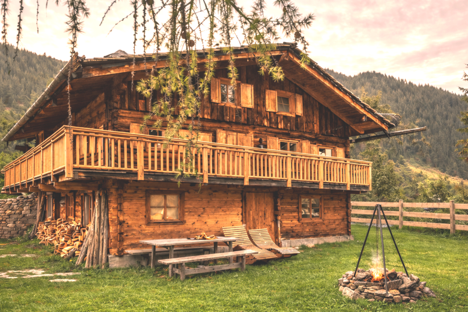 Ein charmantes Holzhaus mit einem Balkon und einem Garten. Im Vordergrund gibt es einen Grillplatz mit einem Lagerfeuer und Blick auf die Berge.