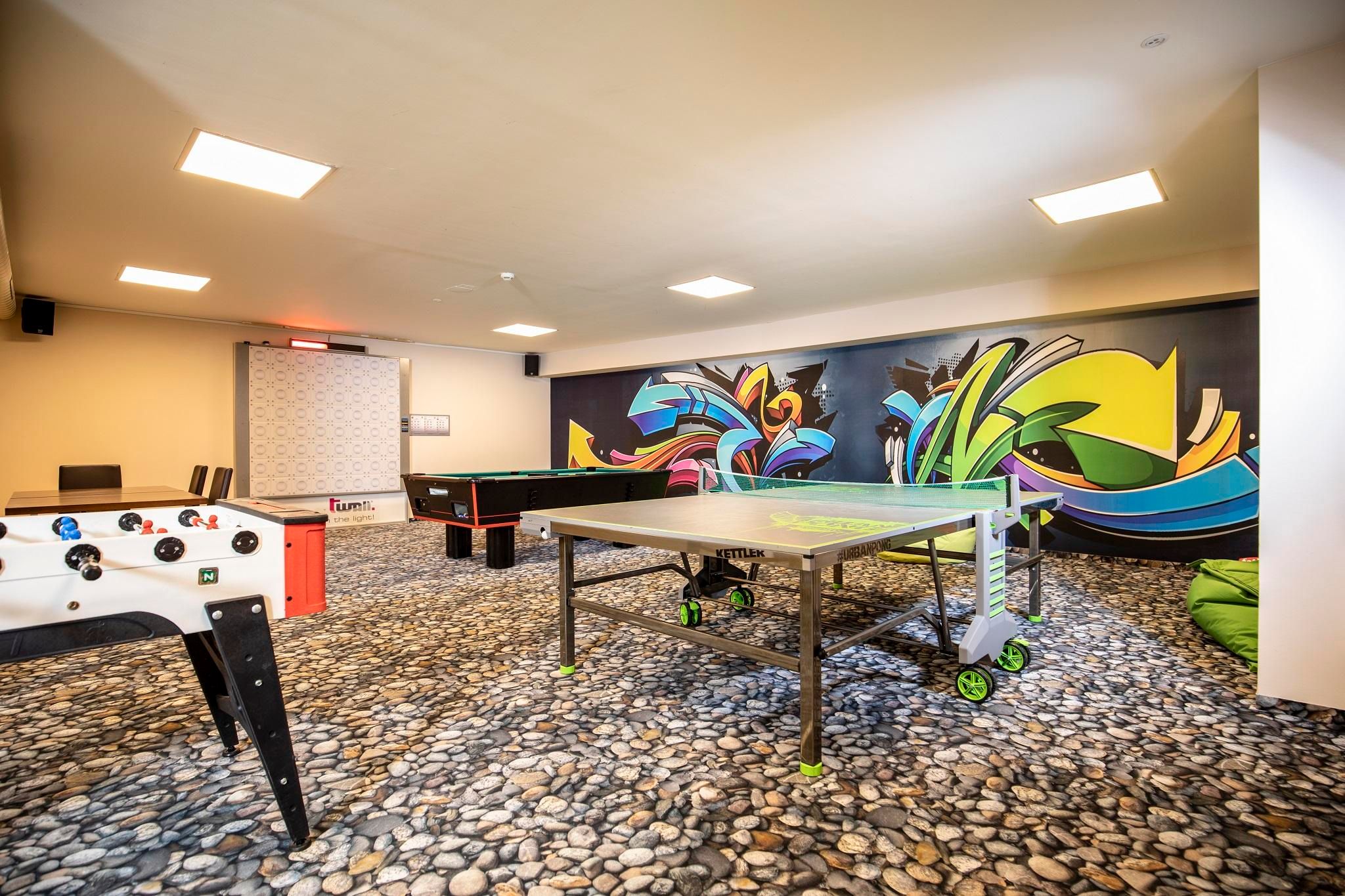 Ein moderner Freizeitraum mit einem Tischtennistisch und einem Tischfußballspiel. Die Wände sind mit bunten Graffiti verziert, und der Boden besteht aus Kieselsteinen.