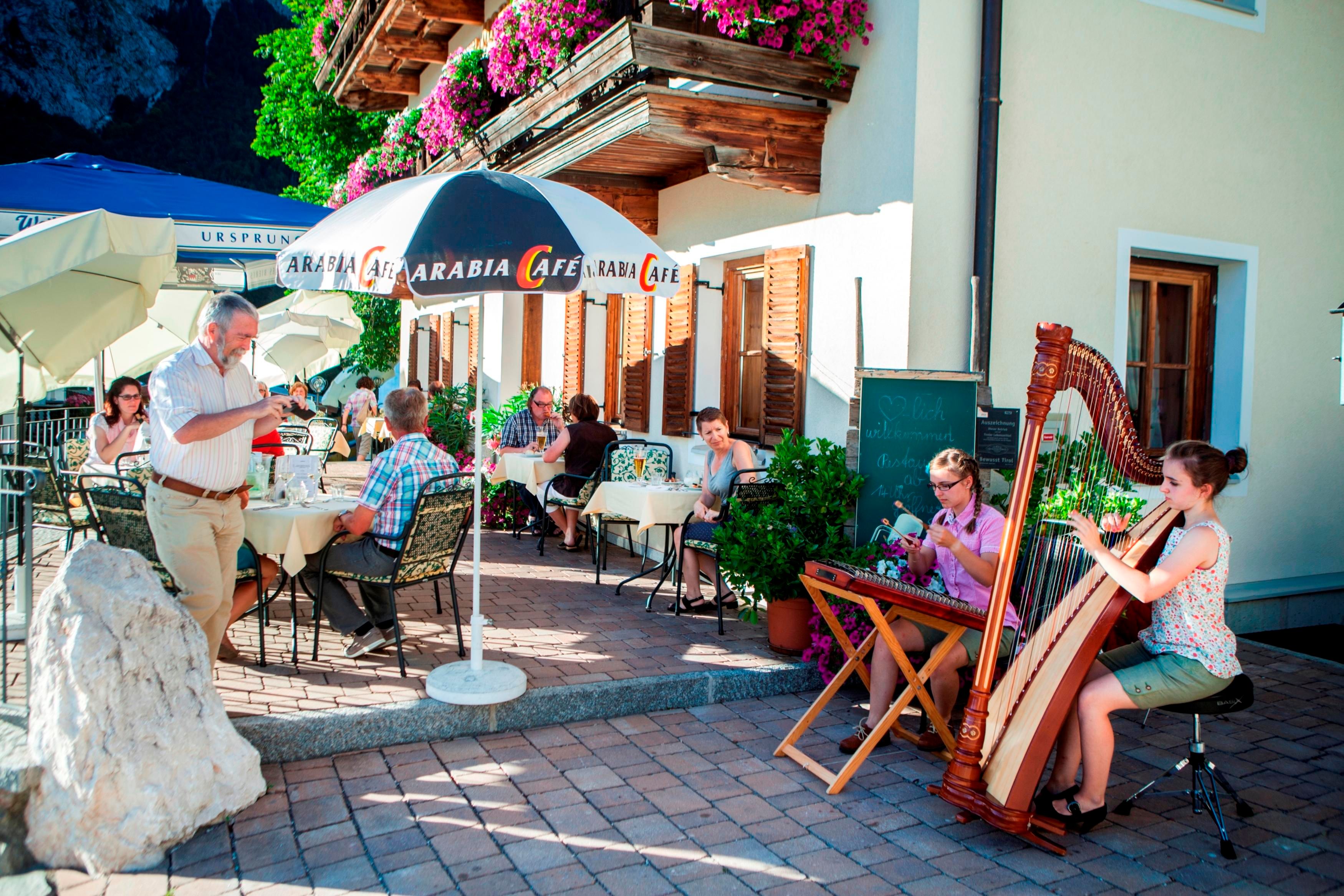 Ein gemütliches Straßencafé mit Gästen, die an Tischen sitzen. Eine Harfenspielerin sorgt für musikalische Unterhaltung.