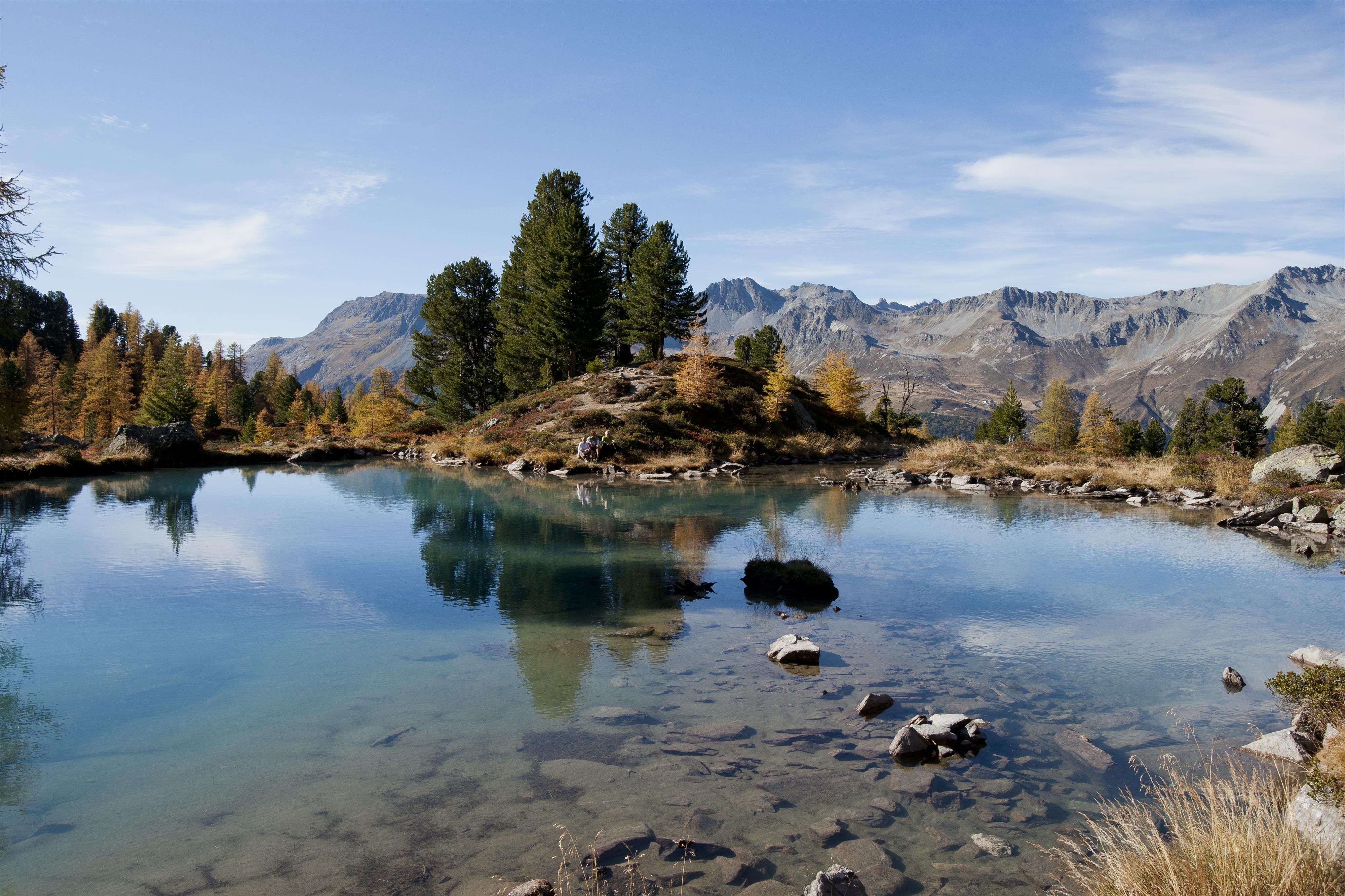Ein klarer Bergsee umgeben von grünen Bäumen und majestätischen Bergen. Die ruhige Wasseroberfläche spiegelt die Umgebung wider.