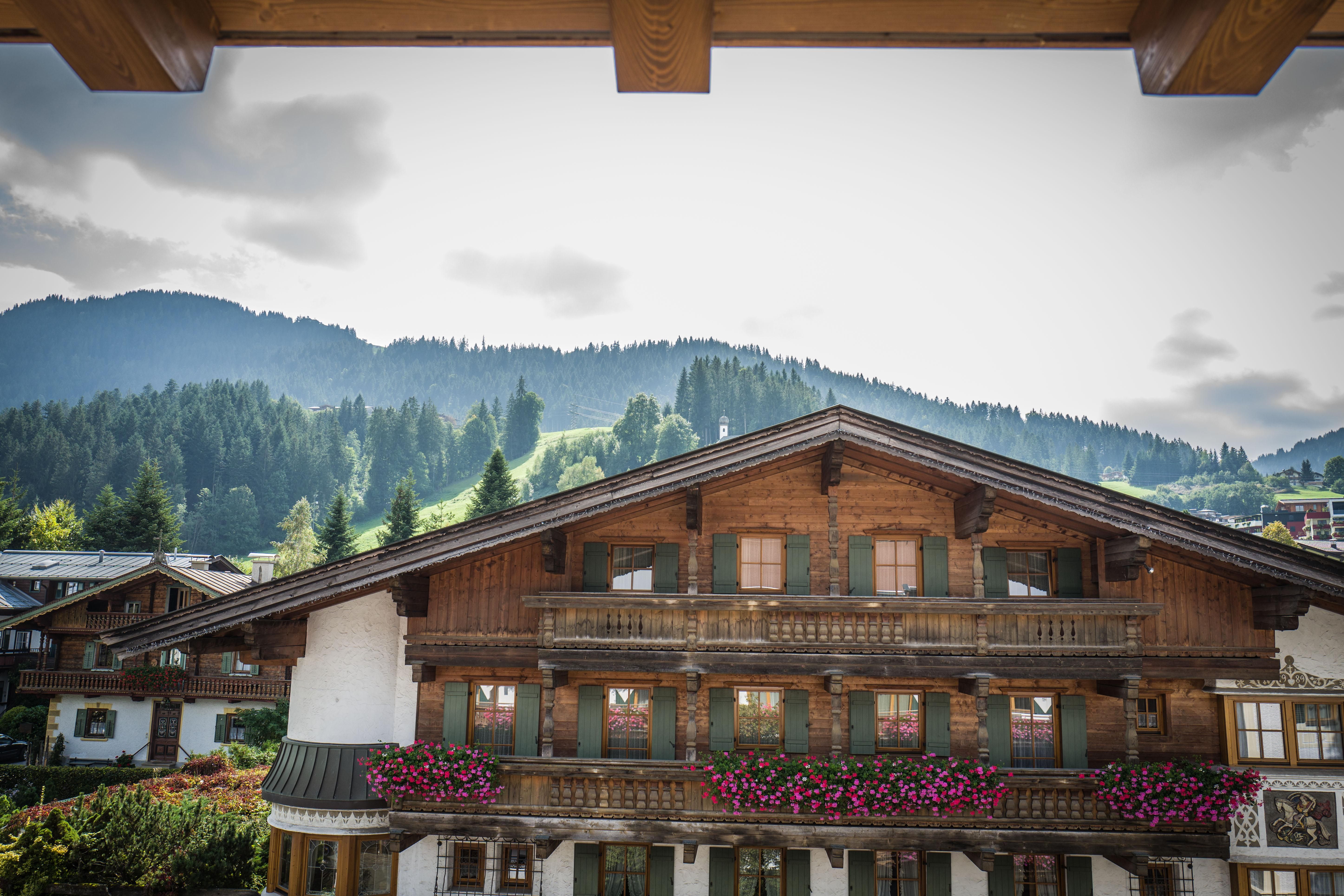 Ein traditionelles Holzhaus mit bunten Blumenbalkonen und Blick auf die Berge. Der Himmel ist bewölkt und die Umgebung ist grün und hügelig.