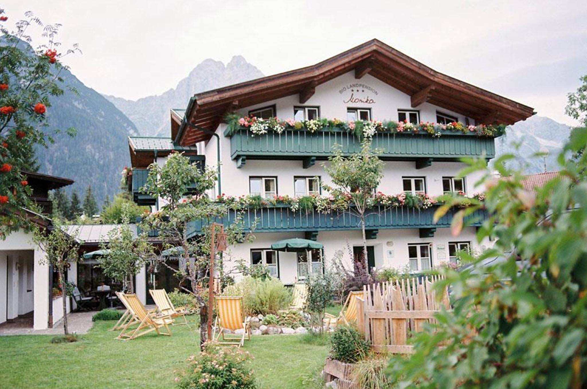 Ein charmantes Haus mit grünen Balkonen und blühenden Pflanzen. Im Vordergrund gibt es einen Garten mit Liegestühlen und alpenländischer Landschaft im Hintergrund.