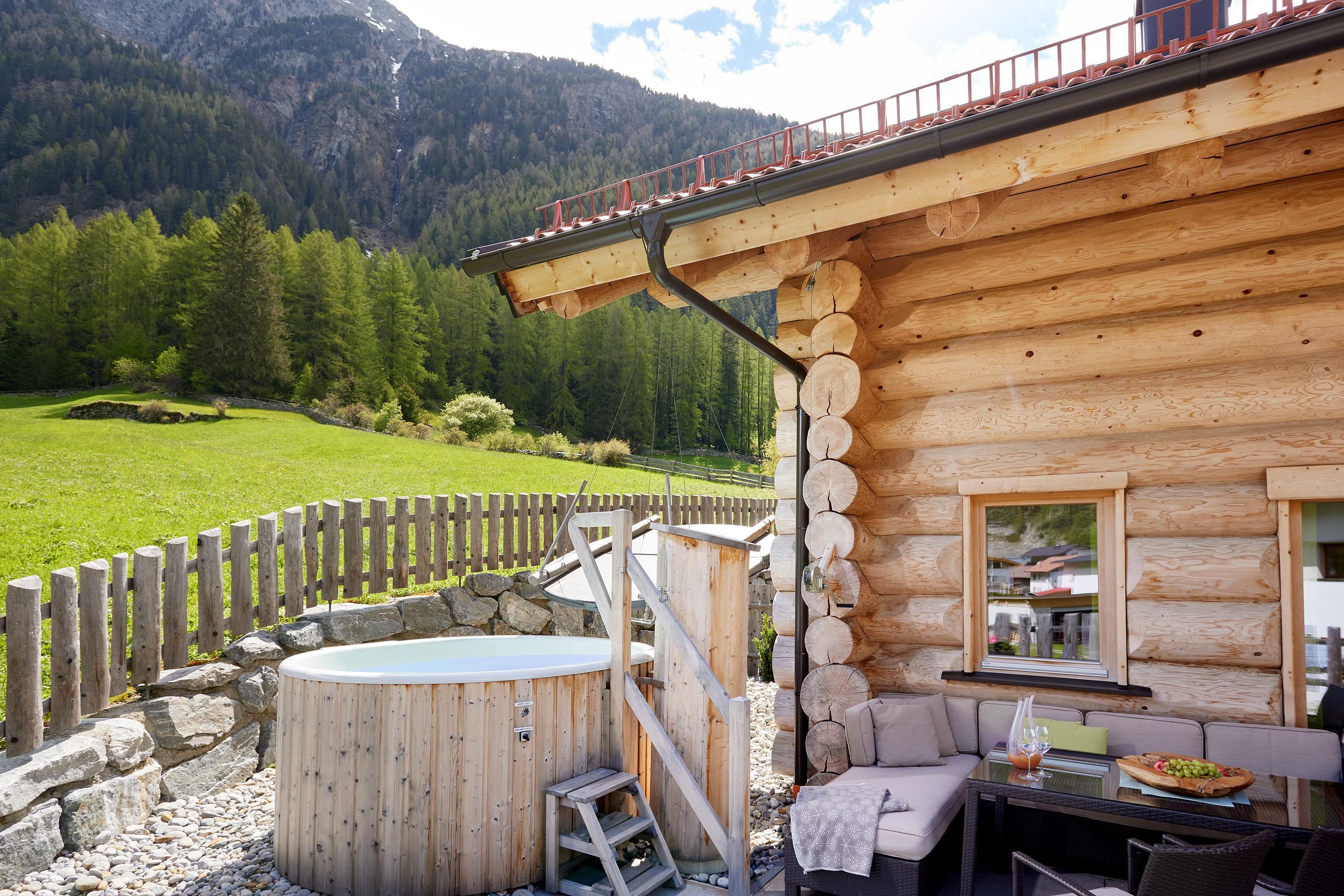 Ein hübsches Holzhaus inmitten von grünen Wiesen und Bergen. Draußen befindet sich ein gemütlicher Jacuzzi.