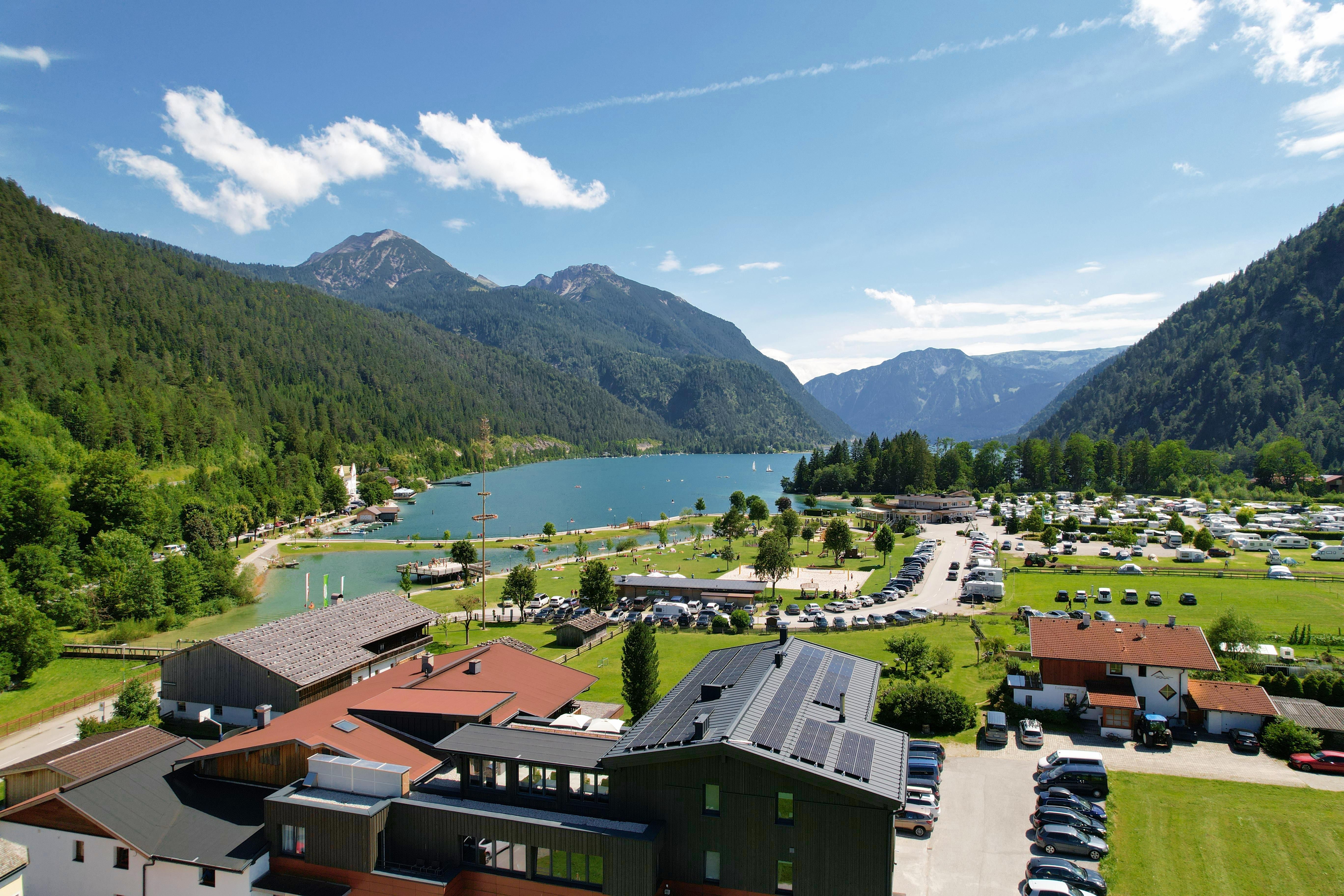 Ein malerischer See umgeben von Bergen und grünen Wäldern. Im Vordergrund sind schöne Gebäude und ein Campingplatz zu sehen.