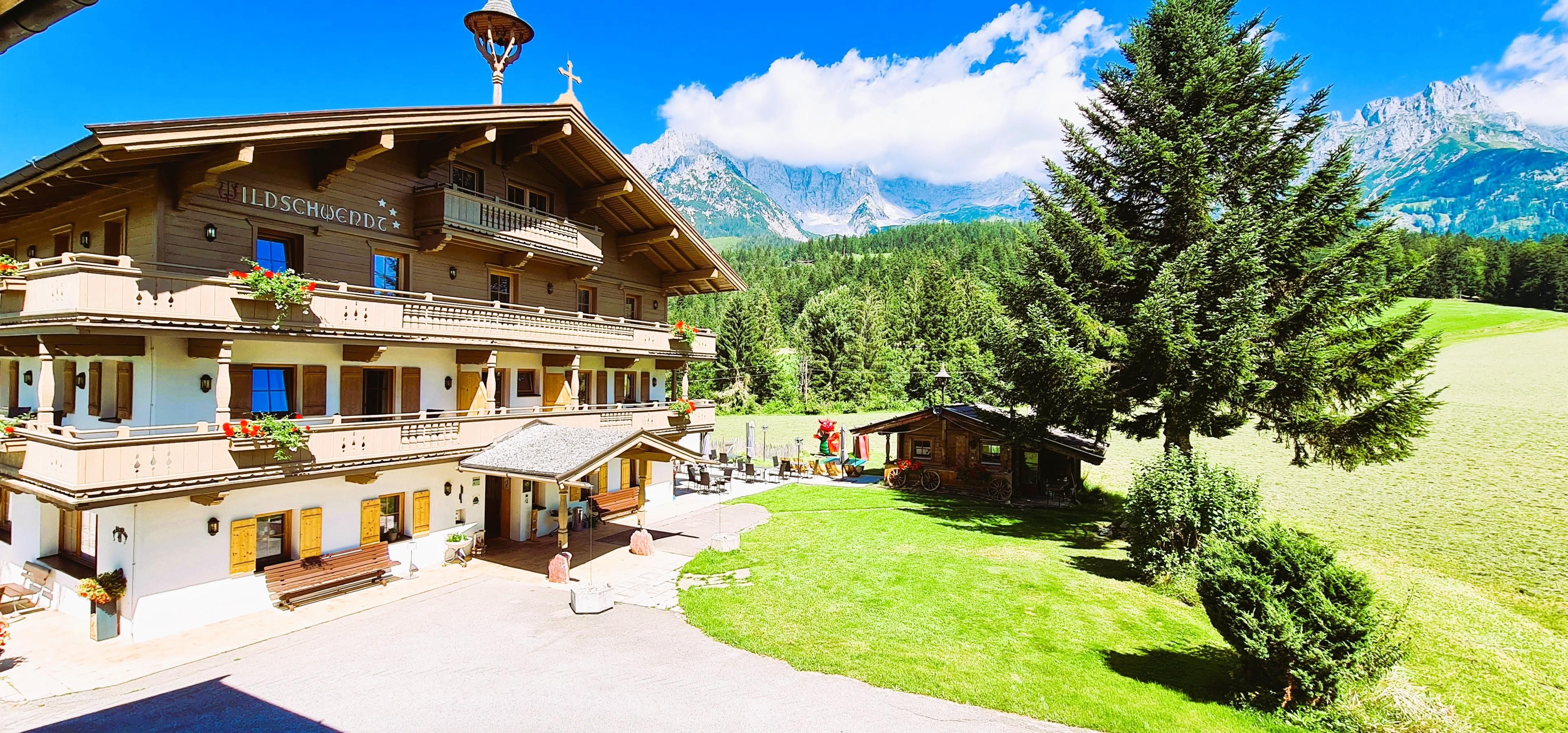 Ein schönes Chalet in den Alpen mit üppigem Grün und hohen Bäumen. Im Hintergrund sind beeindruckende Berge und ein blauer Himmel zu sehen.